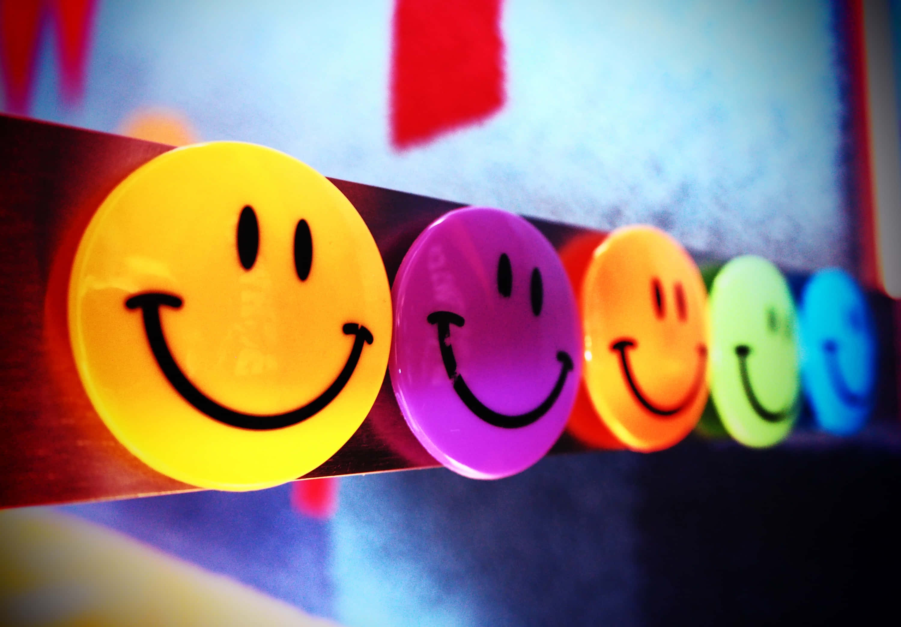 Smiley 桌布 壁紙 — 一排色彩繽紛、有光澤的笑臉按鈕，包括黃色、紫色、橙色、綠色和藍色，排列在深色表面上，背景顏色模糊。