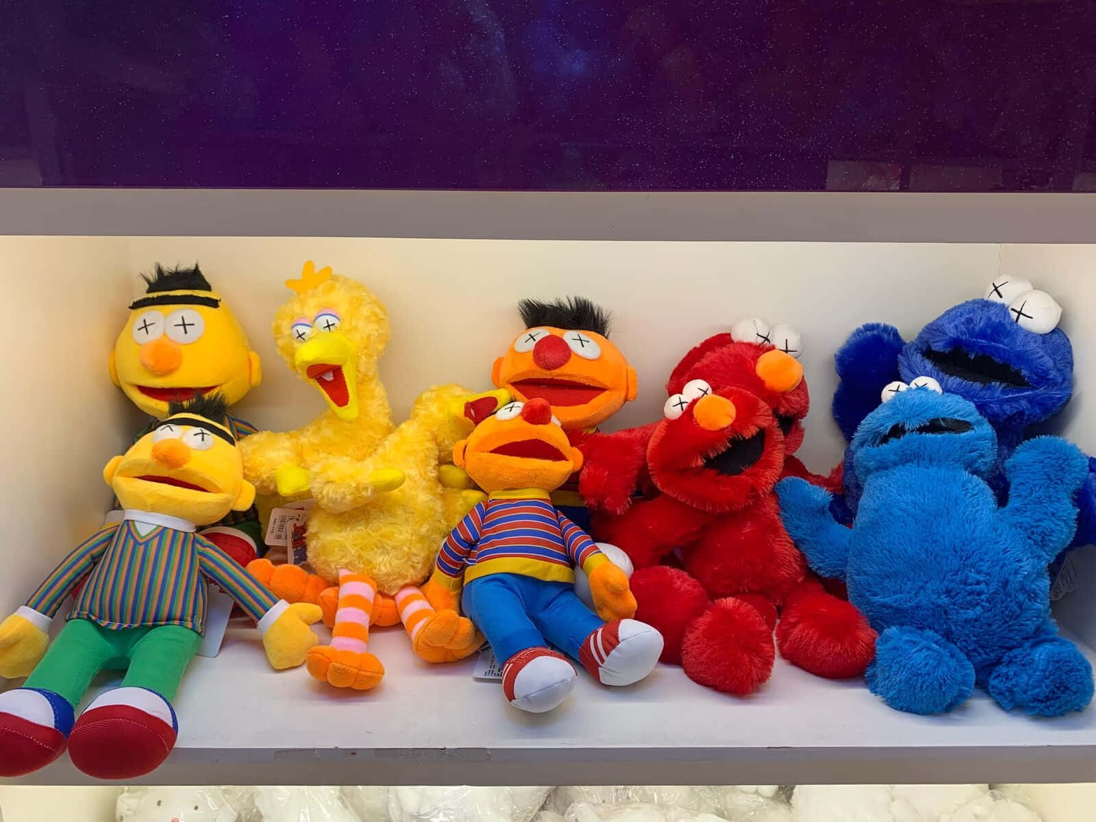 Kaws Sesame Street 桌布 壁紙 — 一排色彩繽紛、眼睛呈X形的芝麻街毛絨玩具放在白色架子上，背景是深紫色牆壁。