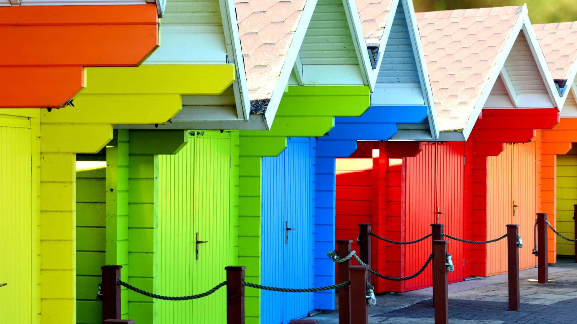Beach Huts 桌布 壁紙 — 一排色彩繽紛的海灘小屋，採用大膽的原色調和瓷磚屋頂，在明亮的日光下顯得生機勃勃。
