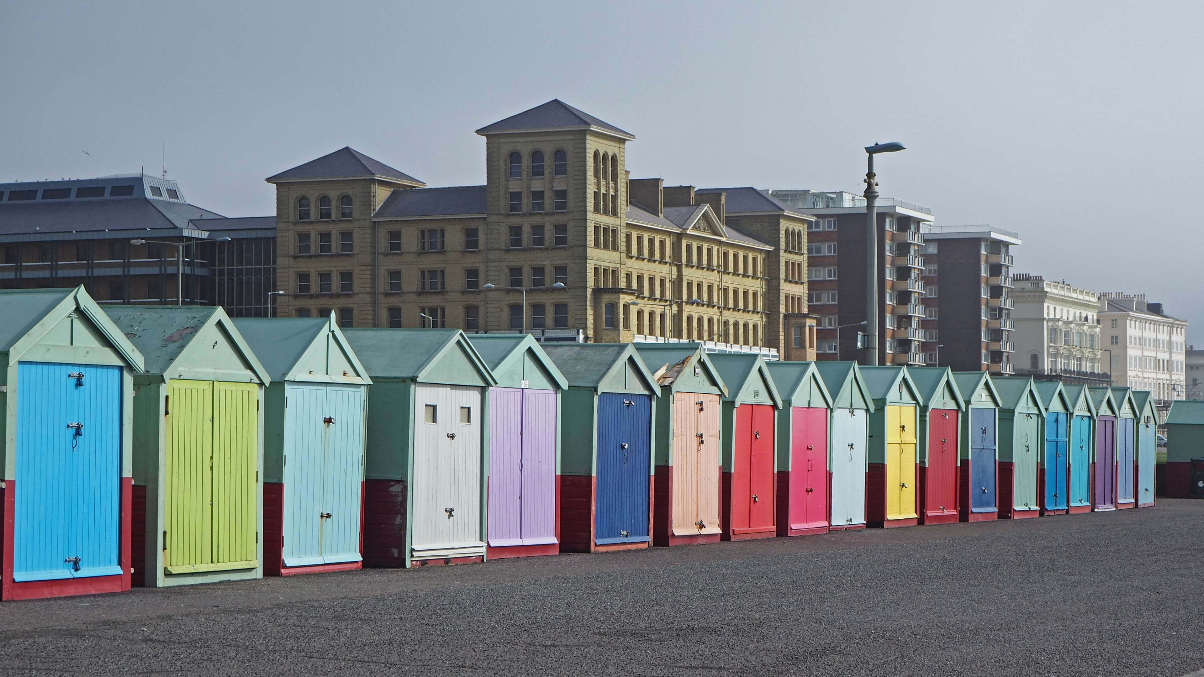 Beach Huts 桌布 壁紙 — 一排色彩鮮艷的沙灘小屋排列在鋪砌的海濱長廊上，背景是歷史建築，天空多雲。