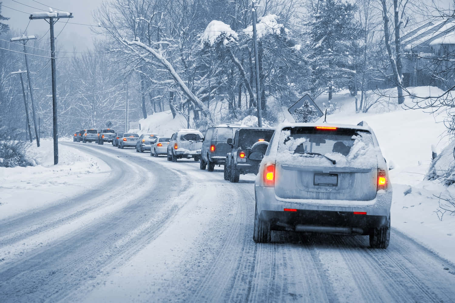 Winter Driving 桌布 壁紙 — 一排被雪覆蓋的汽車在灰色天空下，沿著兩旁有光禿樹木和電線桿的蜿蜒冬季道路緩慢行駛。