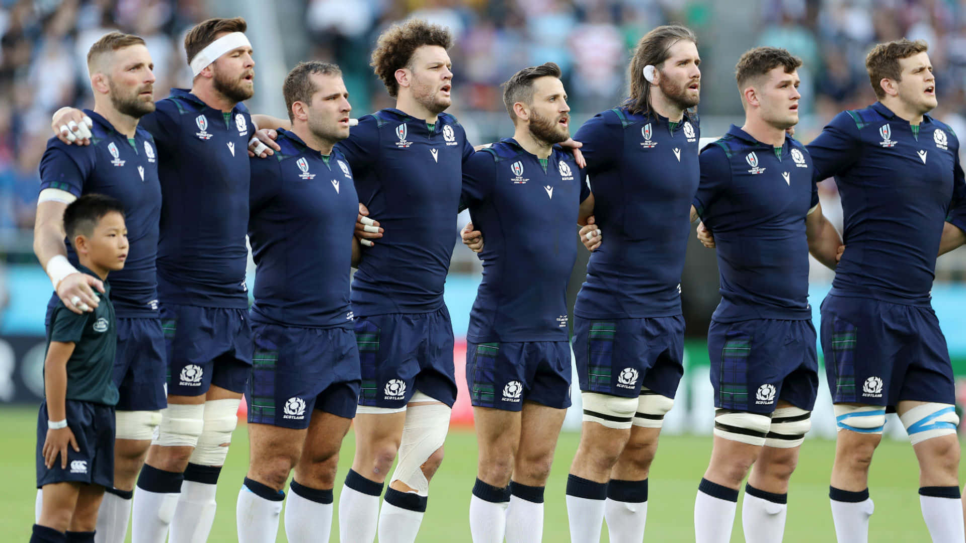Scotland Rugby 桌布 壁紙 — 一排身穿海軍藍球衣的蘇格蘭欖球運動員在國歌奏起時，在綠色球場上莊嚴地互相搭肩站立。