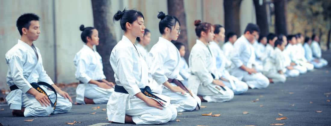 Martial Arts Uniforms 桌布 壁紙 — 一排身穿白色制服的武術家跪在戶外冥想，周圍是落葉的樹木。