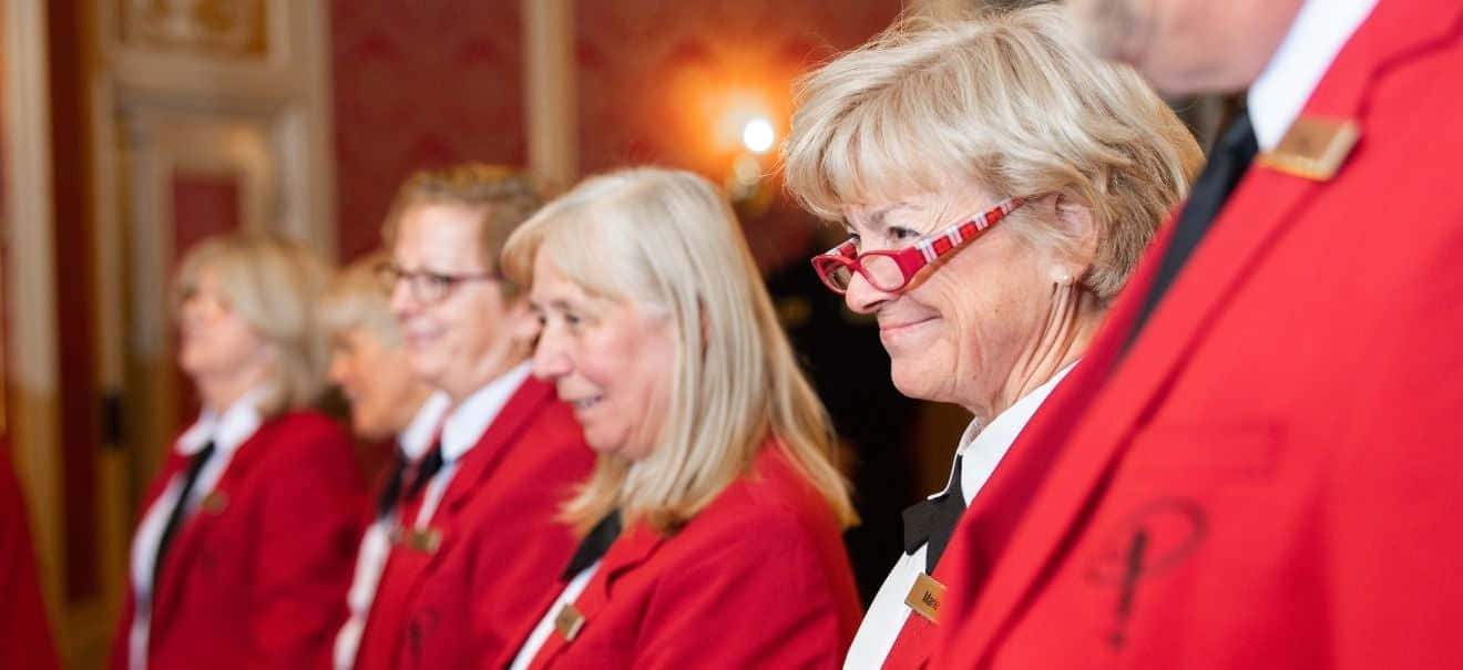 Red Coat 桌布 壁紙 — 一排身穿紅色外套、戴著眼鏡的微笑女性坐在裝飾華麗、燈光溫暖的房間裡。