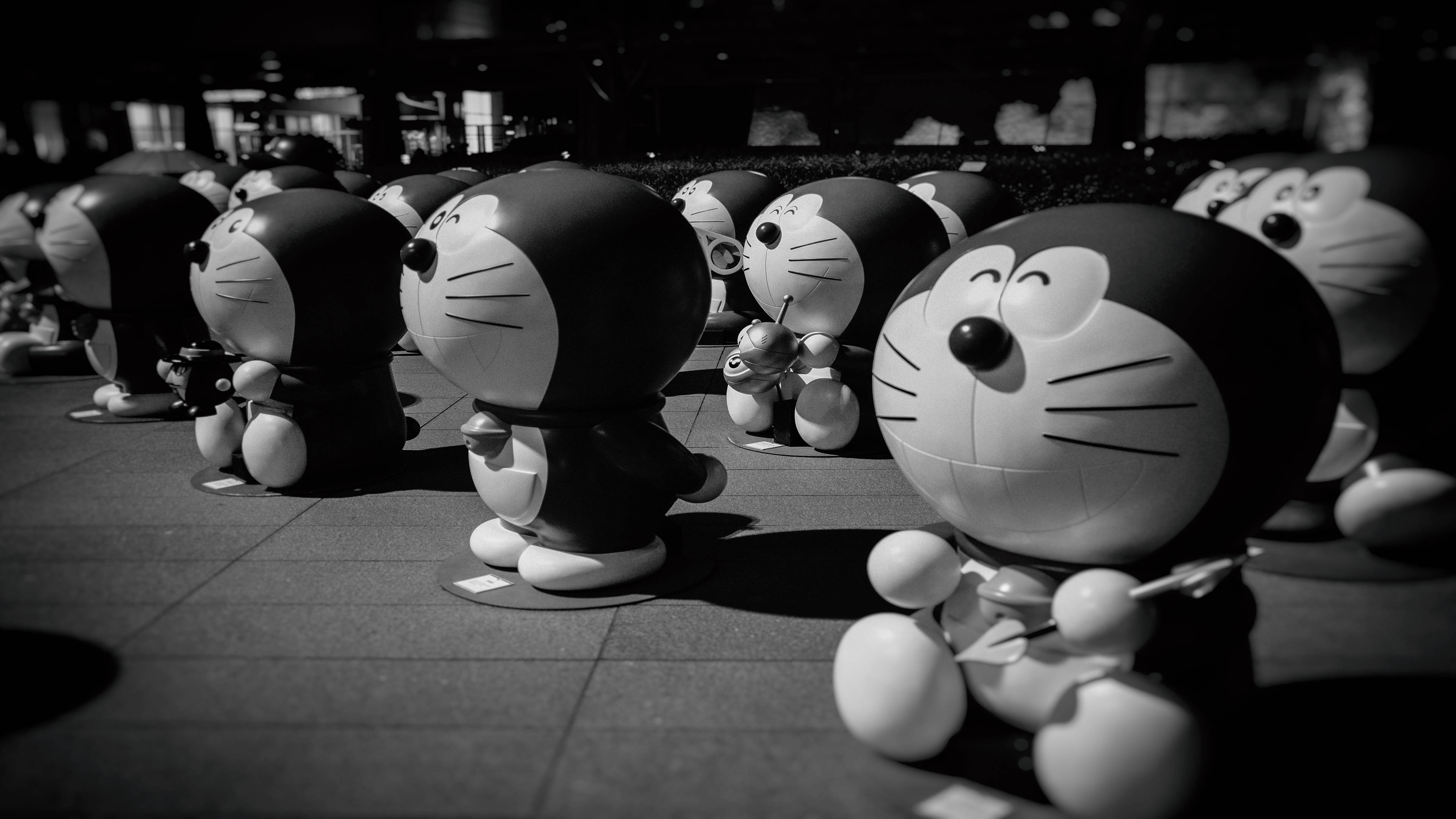 Doraemon 桌布 壁紙 — 一排黑白多啦A夢雕像站在瓷磚廣場上，夜間燈光投射出戲劇性的陰影。