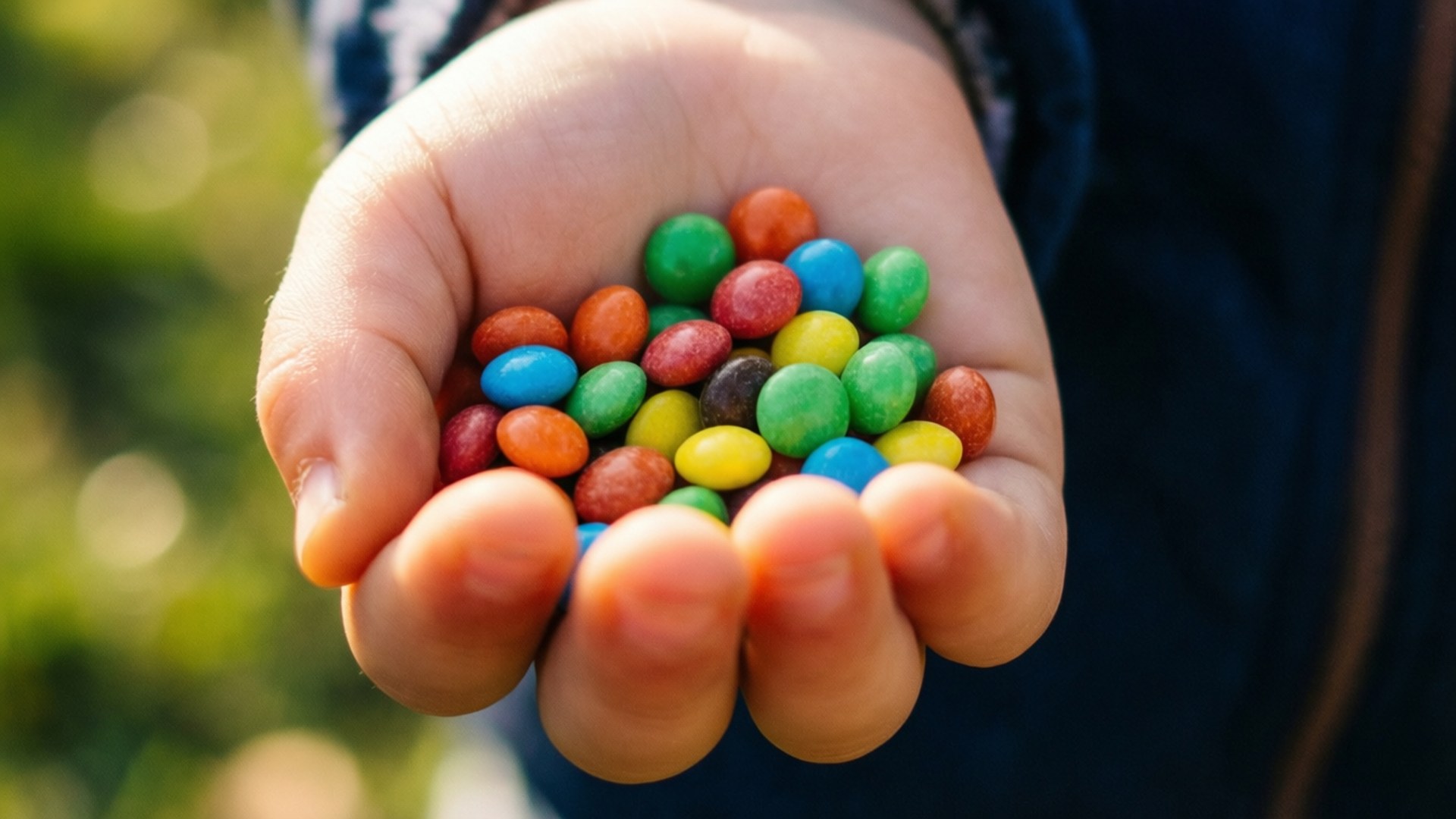 一握Smarties 壁紙 — 孩子張開的手掌捧著一堆色彩繽紛的彩虹M&M's糖果，背景是陽光普照的綠色。