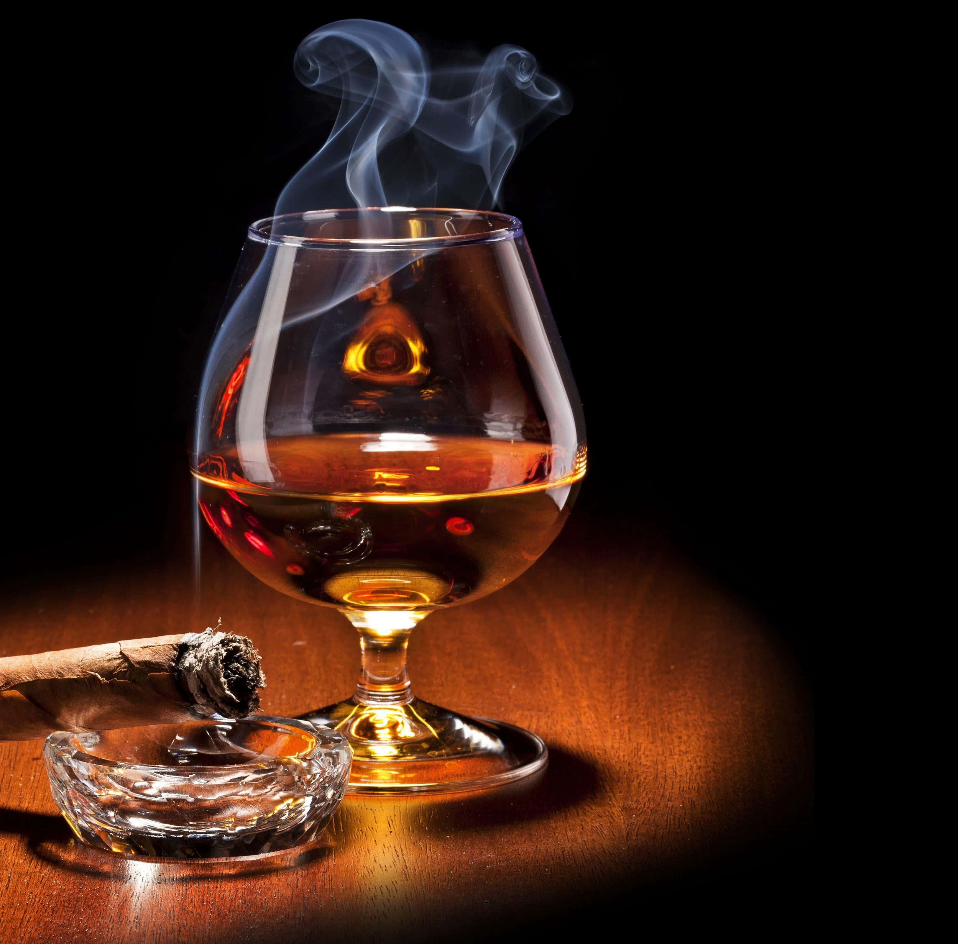 alcohol 桌布 壁紙 — 一支冒煙的雪茄放在煙灰缸裡，旁邊是一杯琥珀色的威士忌，在黑色背景的木桌上溫暖地閃耀。