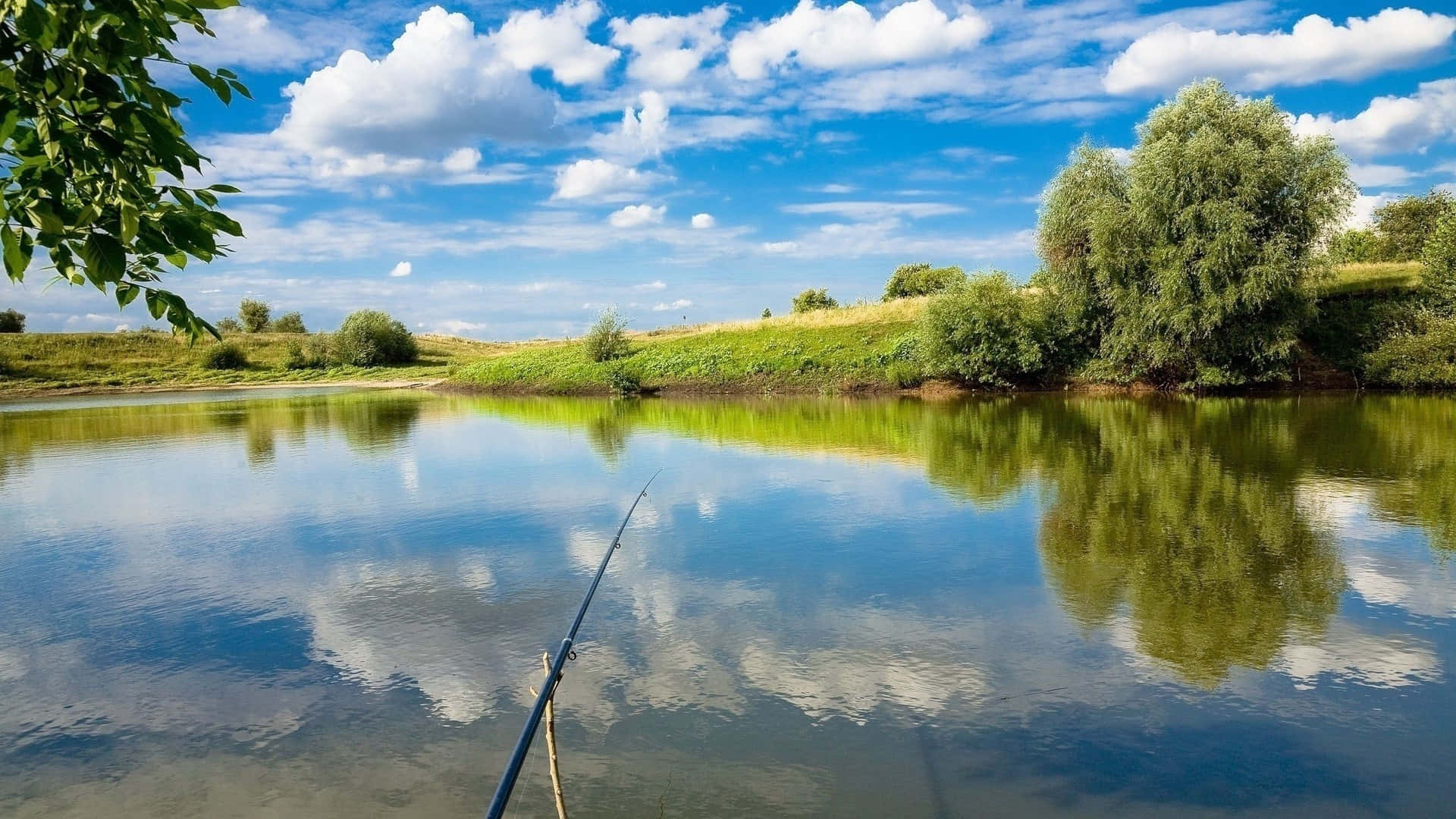 Fishing Rod 桌布 壁紙 — 一支寧靜的魚竿伸展在平靜的河面上，倒映著明亮的藍天、蓬鬆的白雲和茂密的綠樹。