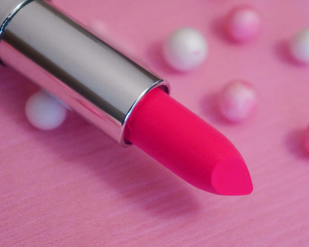 Pink Lipstick 桌布 壁紙 — 一支帶金屬蓋的鮮豔粉紅色唇膏放在柔軟的粉紅色表面上，周圍散落著珍珠。
