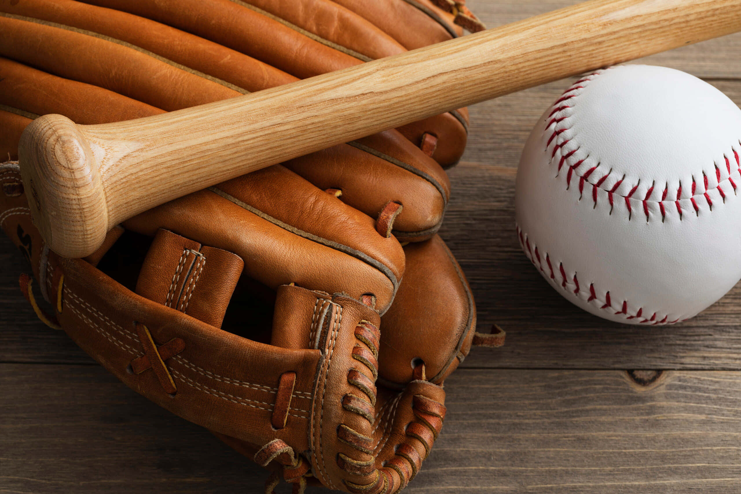 Baseball Gloves 桌布 壁紙 — 一支木製棒球棒放在一個磨損的皮革手套上，旁邊有一個帶紅色縫線的白色棒球，置於質樸的木質表面上。