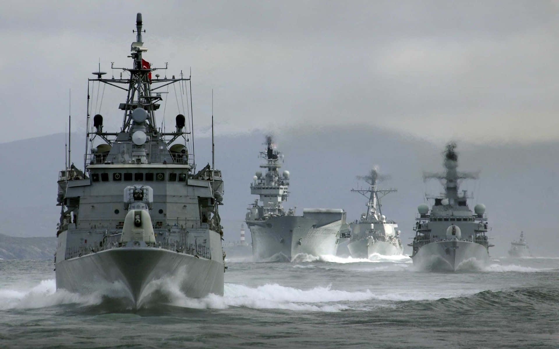 Navy 桌布 壁紙 — 一支灰色軍艦艦隊喺多雲嘅天空下，背景有遠處嘅山脈，穿越波濤洶湧嘅水域。