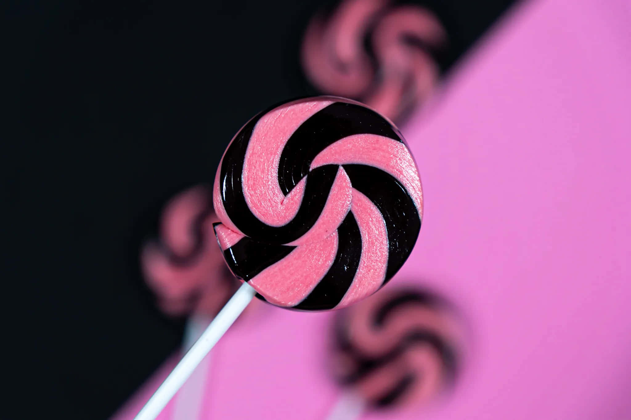 Pink Candy 桌布 壁紙 — 一支白色棒上的光澤粉紅和黑色漩渦棒棒糖，在鮮豔的粉紅和黑色背景前清晰對焦。