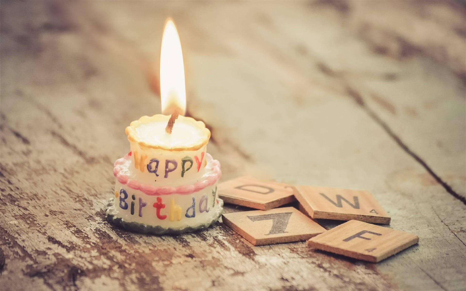 Birthday 桌布 壁紙 — 一支細小的生日蠟燭，上面有 'Happy Birthday' 字樣，在質樸的木桌上明亮地燃燒，旁邊散落著 Scrabble 字母磚。
