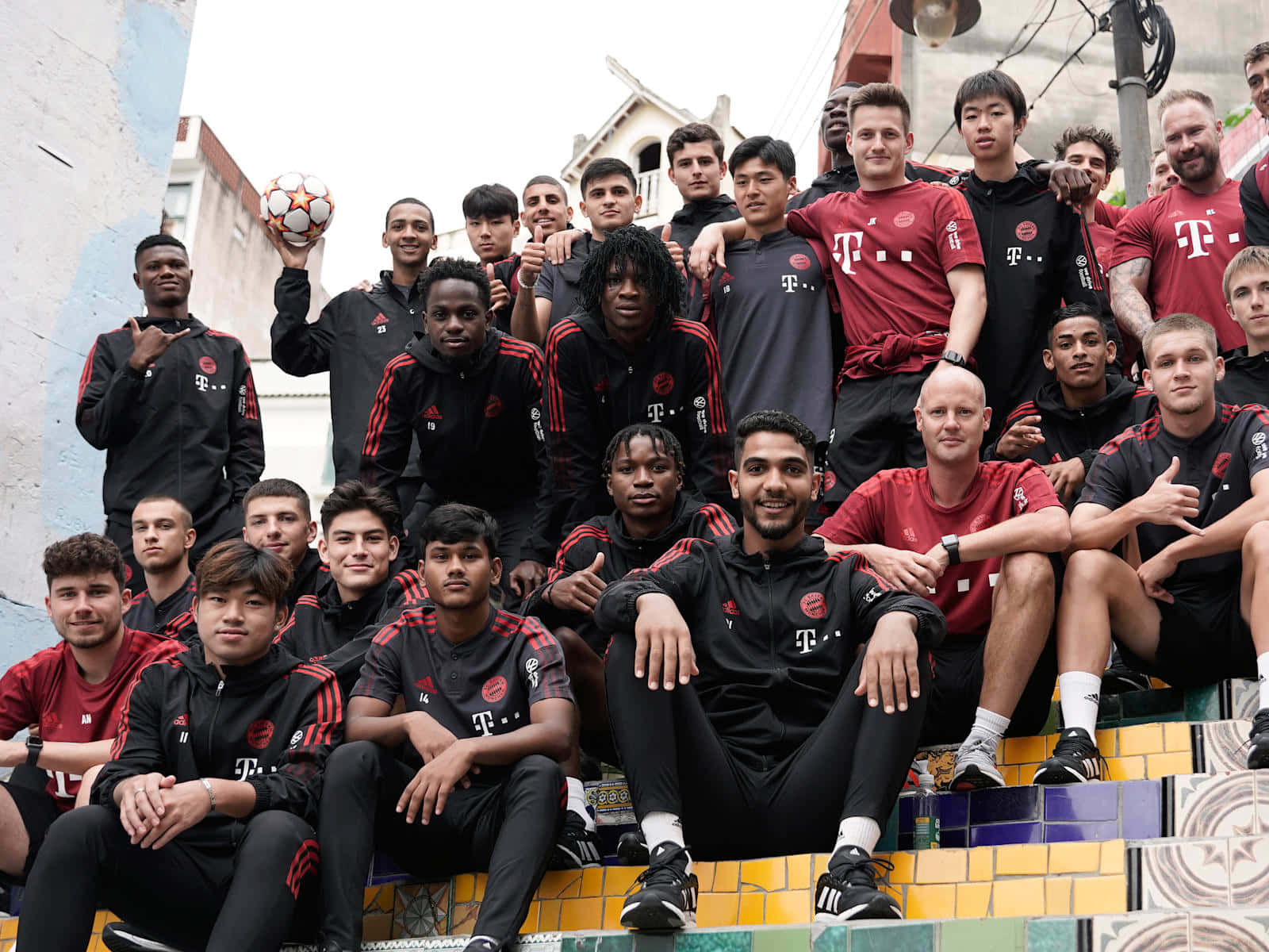 squad 桌布 壁紙 — 一支足球隊穿著紅黑色訓練裝備，在色彩繽紛的瓷磚階梯上一起擺姿勢，其中一名球員手持足球。