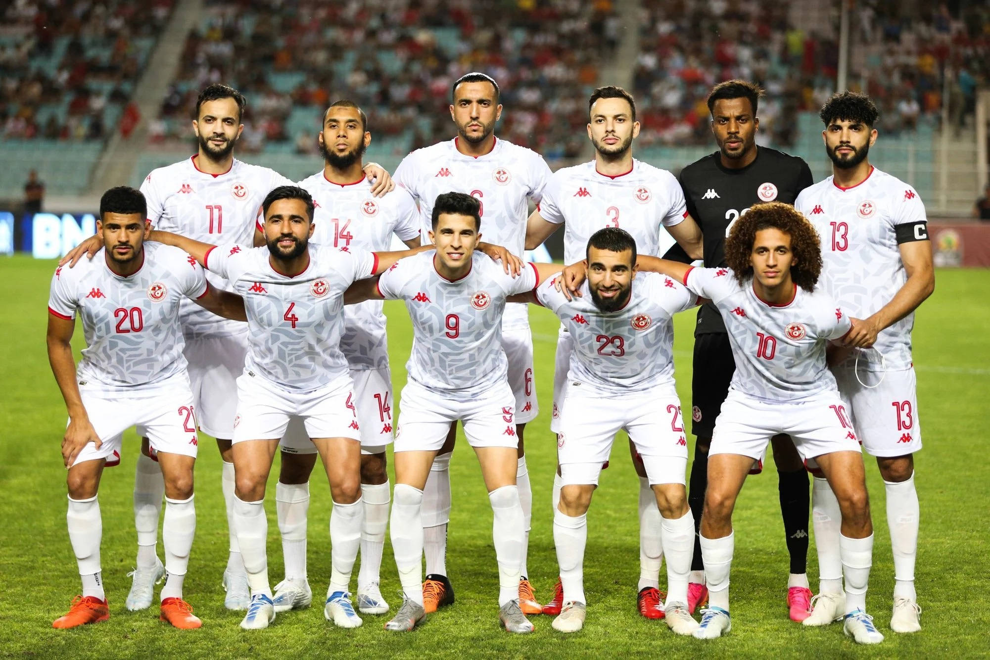 Tunisia 桌布 壁紙 — 一支身穿白色球衣的足球隊在體育館燈光下，於充滿活力的綠色球場上自信地擺姿勢，背景有球迷。