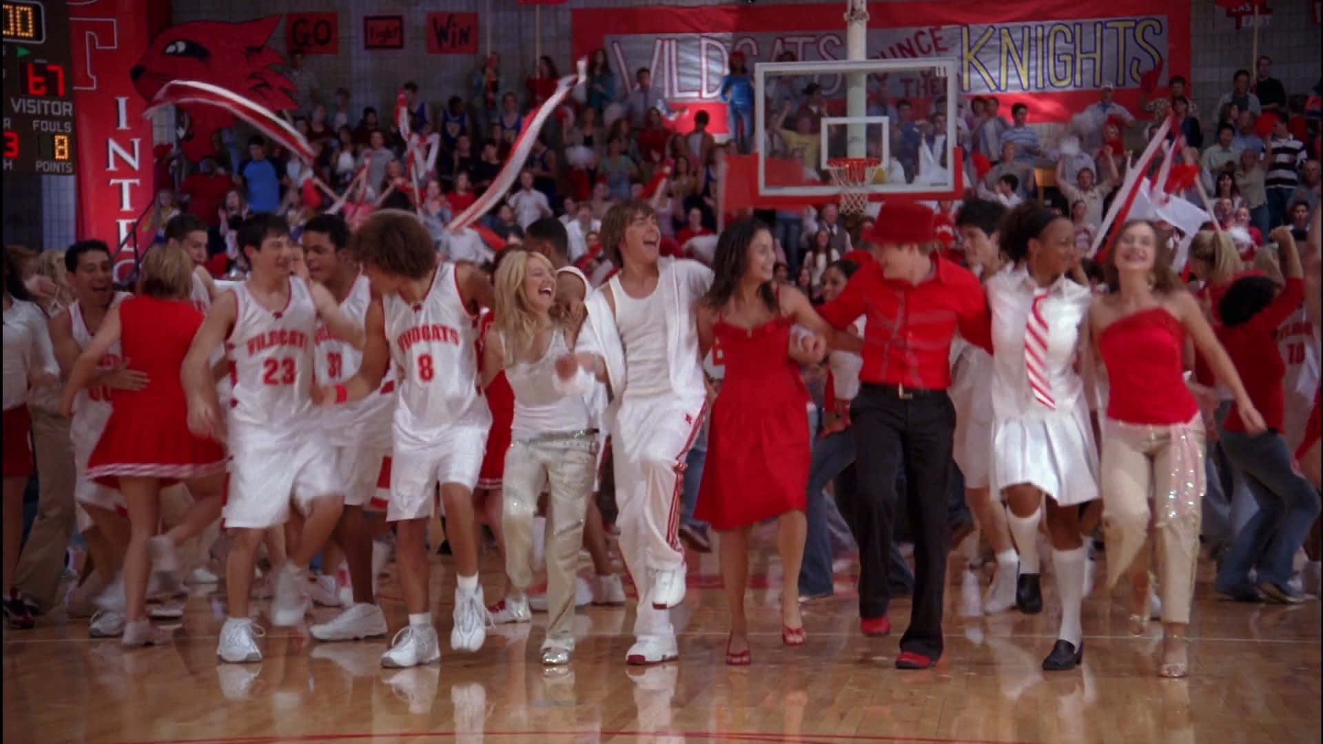 High School Musical 桌布 壁紙 — 一支身穿紅白球衣的籃球隊和啦啦隊在光亮的體育館球場上歡慶勝利。