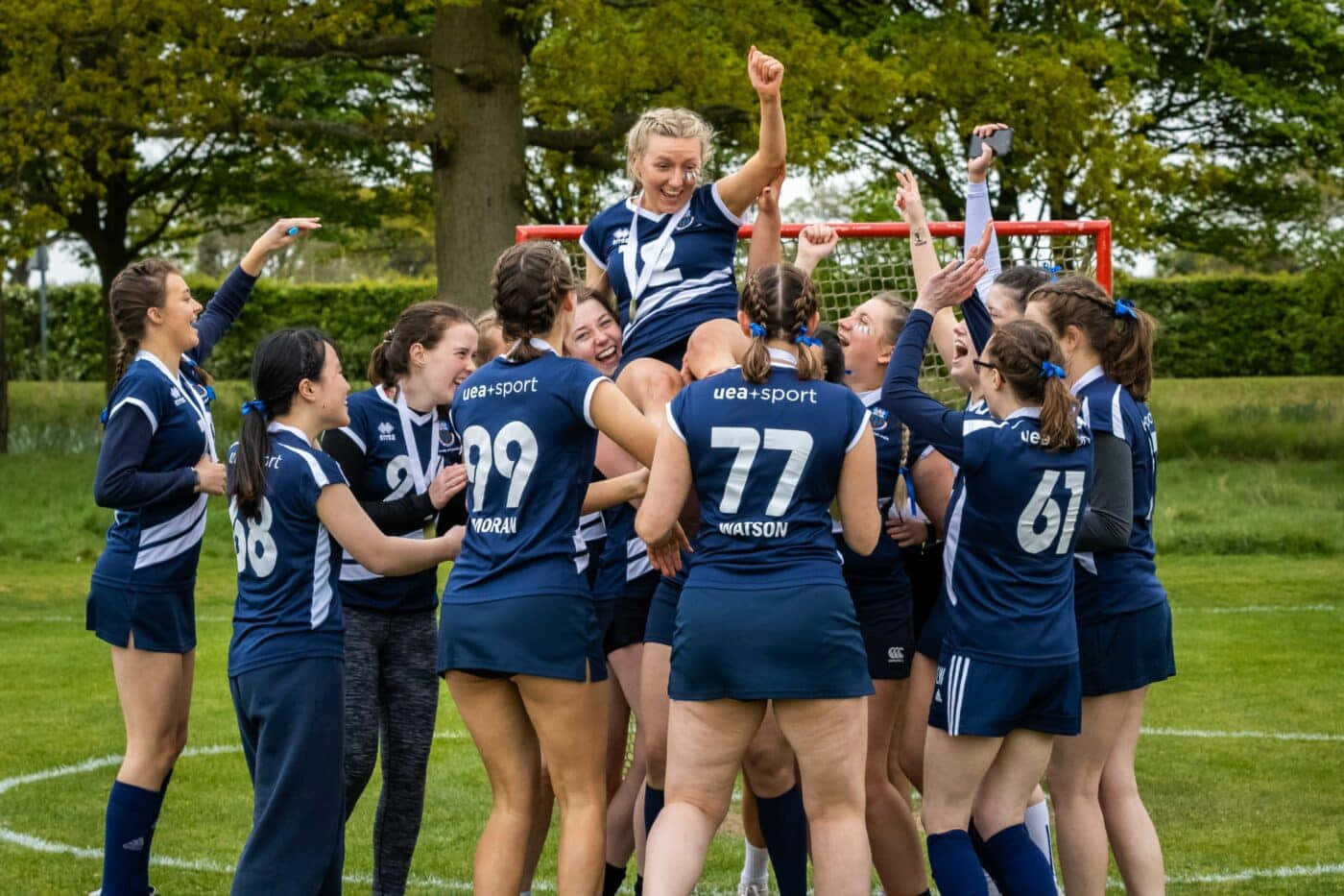 sport 桌布 壁紙 — 一支身穿藍色球衣的女子籃網球隊在綠色的戶外球場上慶祝勝利，周圍樹木環繞。