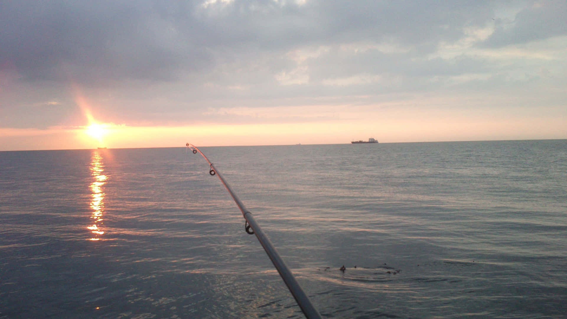 Fishing Rod 桌布 壁紙 — 一支魚竿伸向平靜的海面，太陽在雲層後落下，投射出金色的倒影。