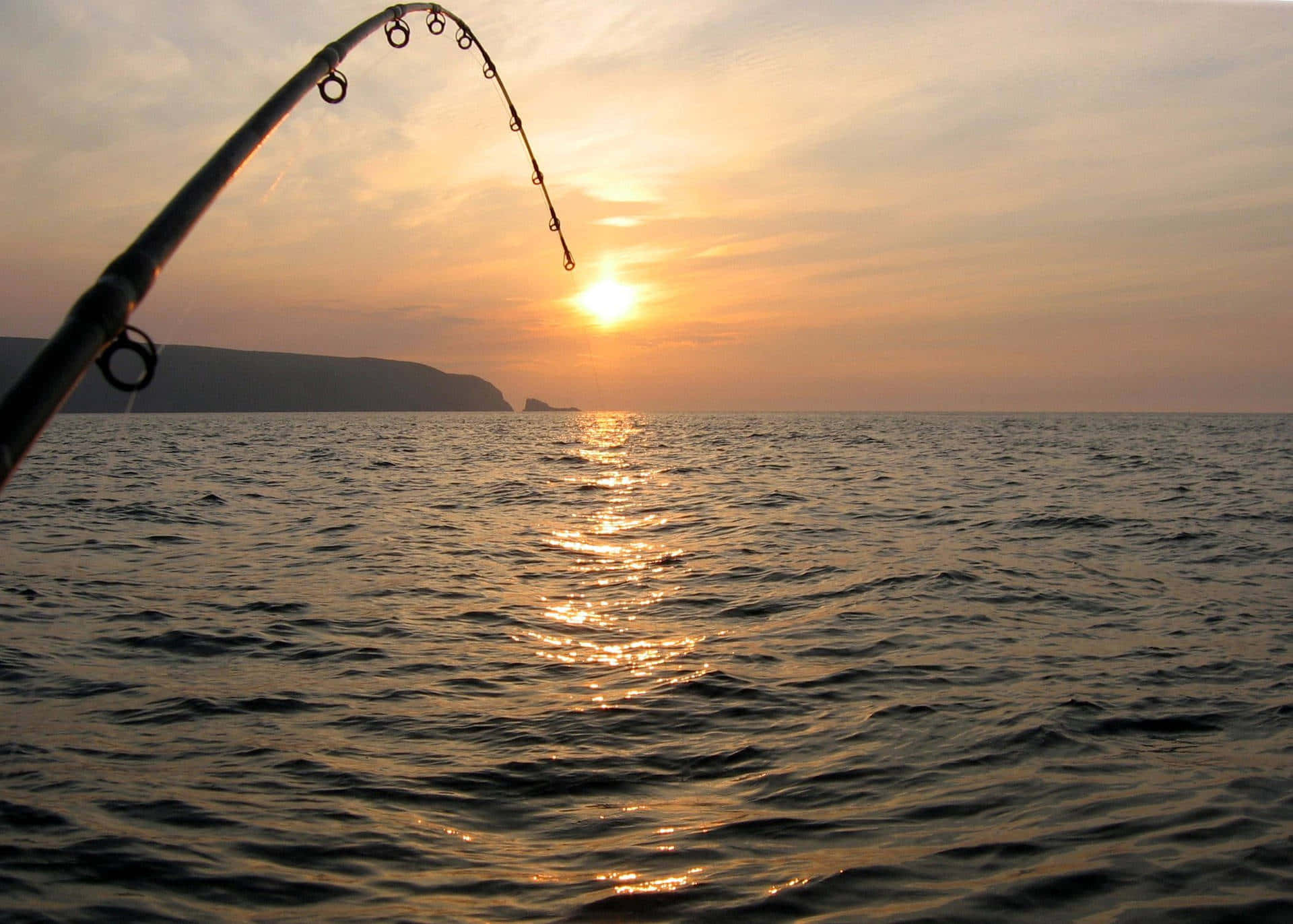 Fishing Bait 桌布 壁紙 — 一支魚竿在平靜的海面上，對著遠方懸崖的金色夕陽彎曲。