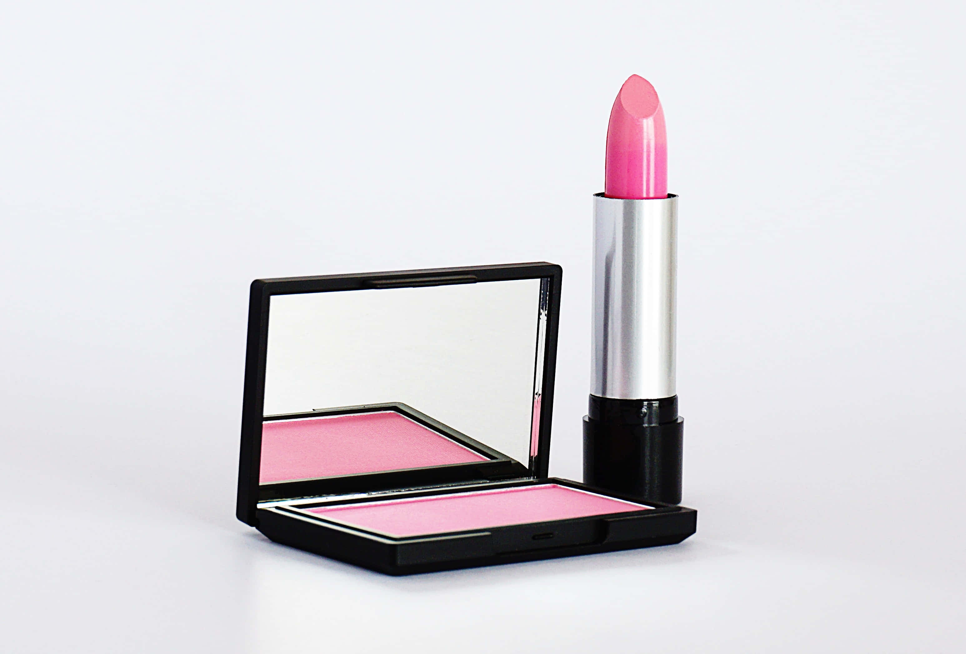Pink Lipstick 桌布 壁紙 — 一支鮮粉紅色唇膏放在一個打開的黑色粉盒旁邊，盒內有相配的胭脂和鏡子，置於潔白表面上。