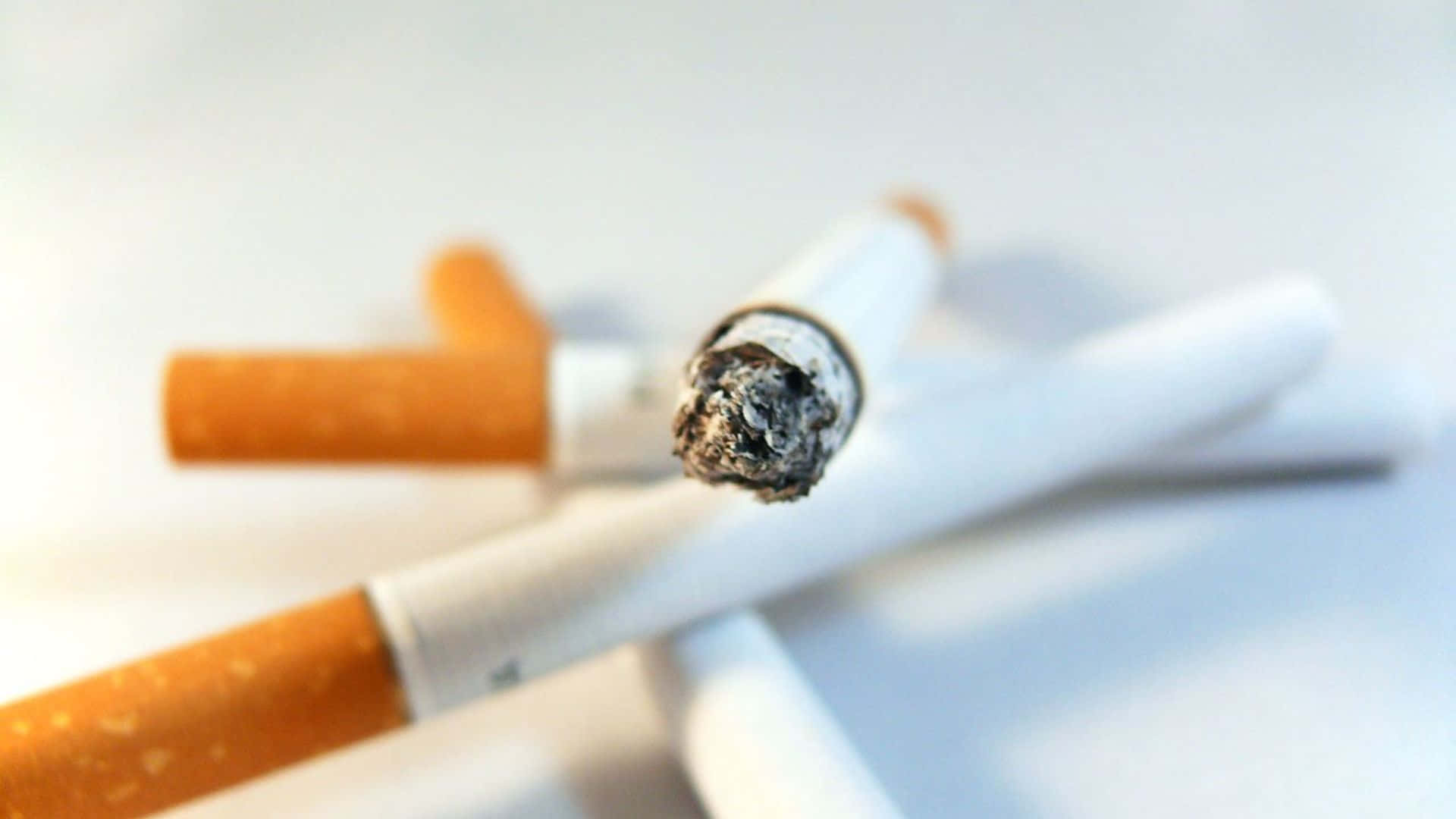 吸煙 桌布 壁紙 — 一支點燃的香煙特寫，帶有灰燼和煙霧，背景為模糊的白色。