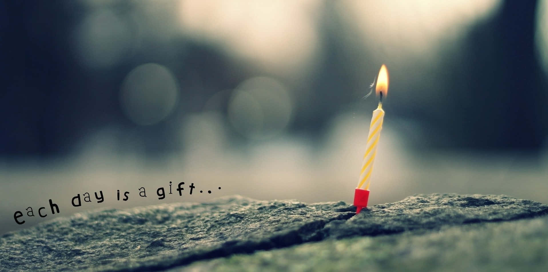 動力 桌布 壁紙 — 一支點燃的黃色條紋生日蠟燭立在粗糙的石頭上，在柔和模糊的背景中溫暖地發光，並附有文字每一天都是禮物。