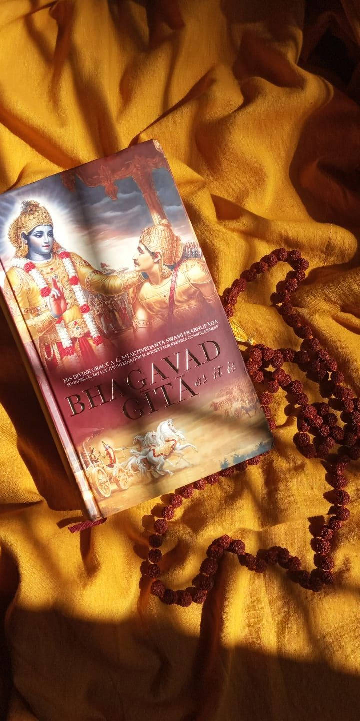 Indian 桌布 壁紙 — 一本《薄伽梵歌》書籍放在金色布料上，旁邊有一串紅色念珠，在溫暖的陽光下。