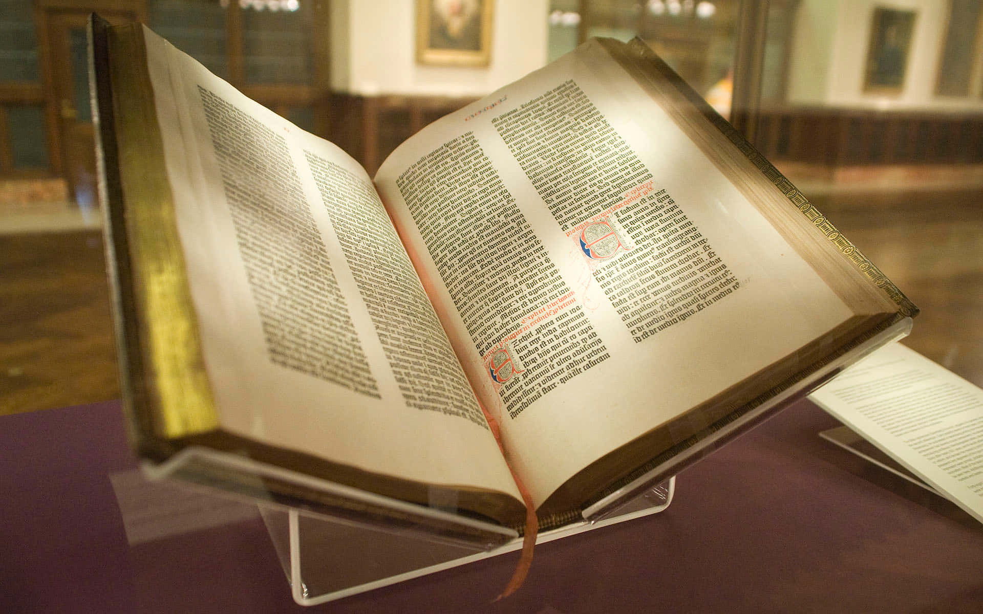 Biblical 桌布 壁紙 — 一本古老嘅彩繪手稿書，打開展示喺玻璃櫃入面，有金色邊緣同紅色墨水嘅首字母。