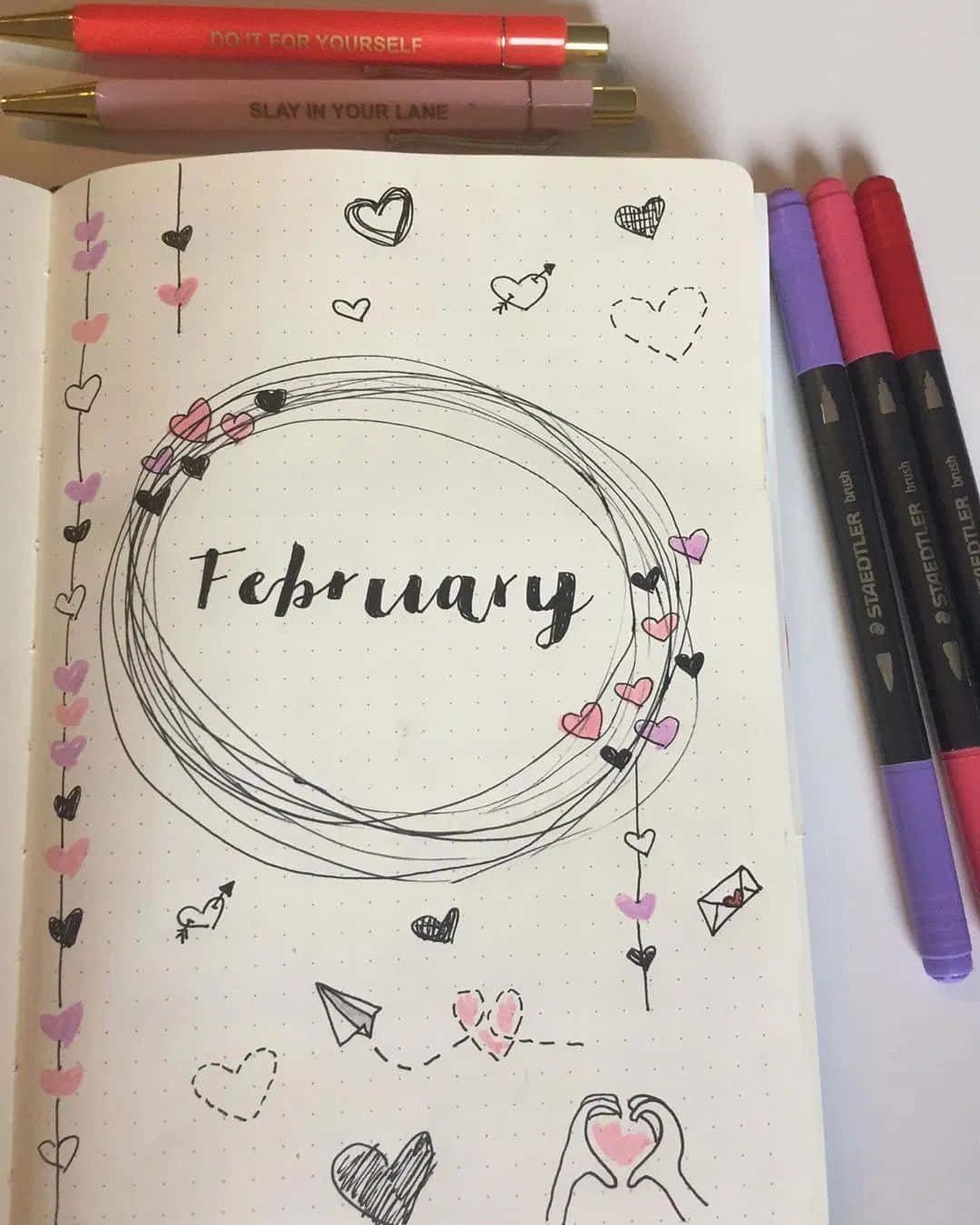 February iPhone 桌布 壁紙 — 一本子彈筆記頁面，二月寫在圓形框架內，裝飾有粉紅色和黑色的心形及塗鴉，周圍環繞著彩色筆。