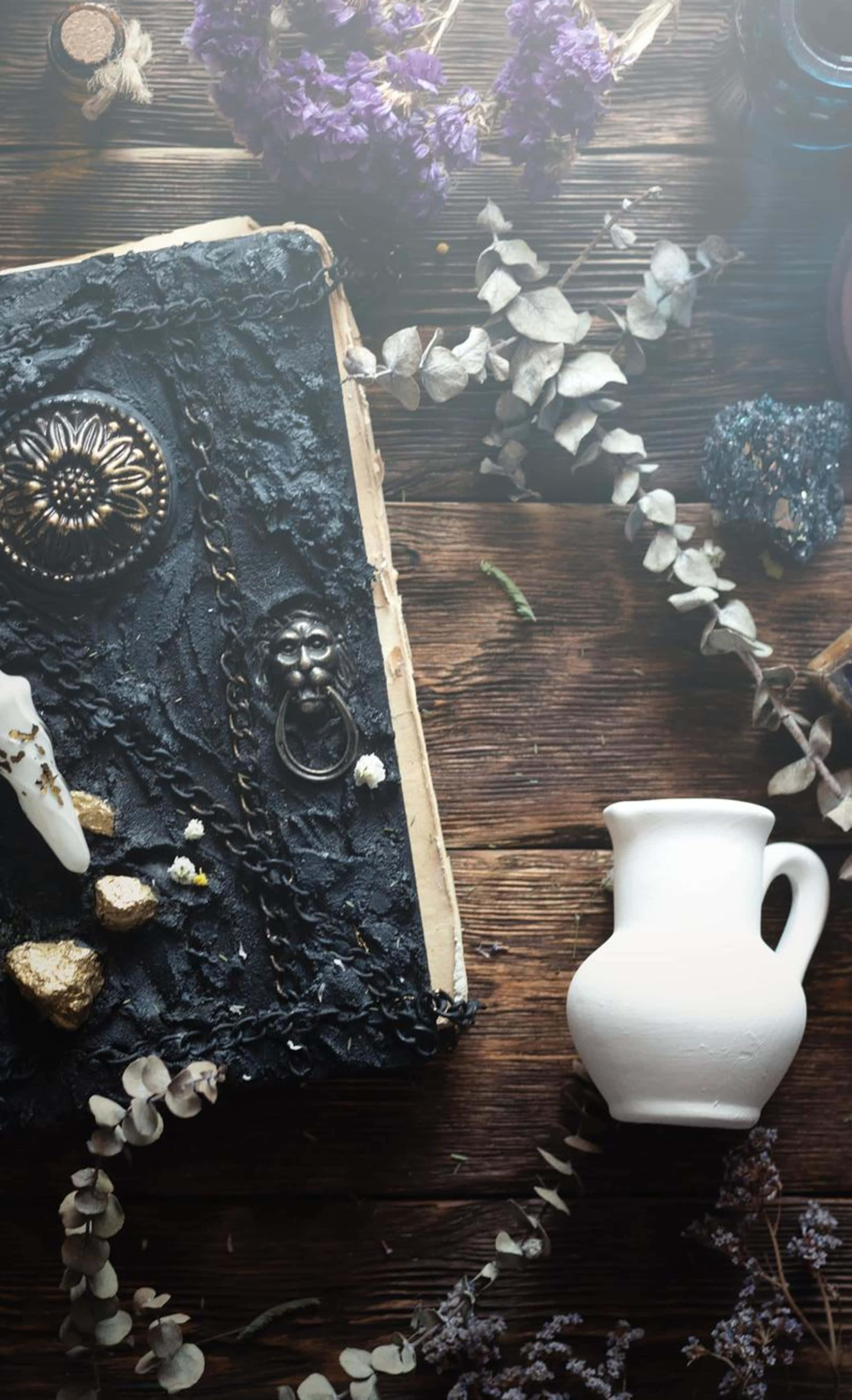 Grimoire 桌布 壁紙 — 一本帶有華麗金屬扣環的古老黑色紋理書本，放在一張深色木桌上，周圍環繞著乾燥的薰衣草桉樹和一個白色陶瓷壺。