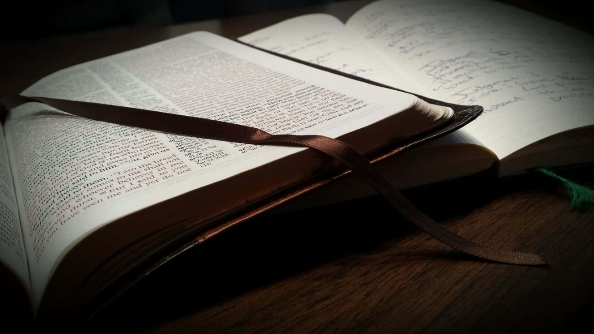 Bible Study 桌布 壁紙 — 一本打開的書，內有手寫筆記和絲帶書籤，放在木質表面上，在溫暖聚焦的光線下。