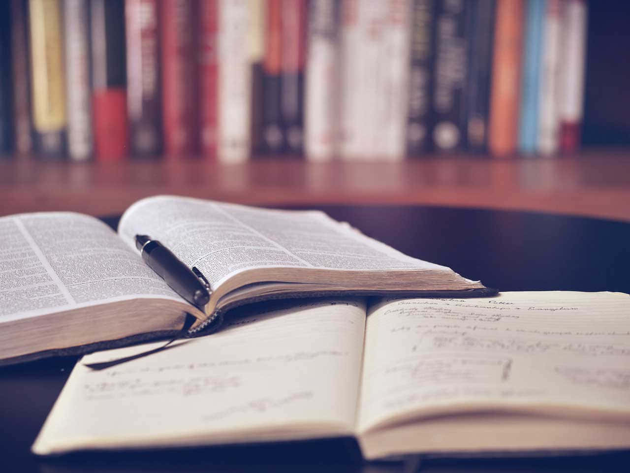 閱讀 桌布 壁紙 — 一本打開的書本連同一支筆放在一本滿是手寫筆記的筆記本上，前面是模糊的書架。