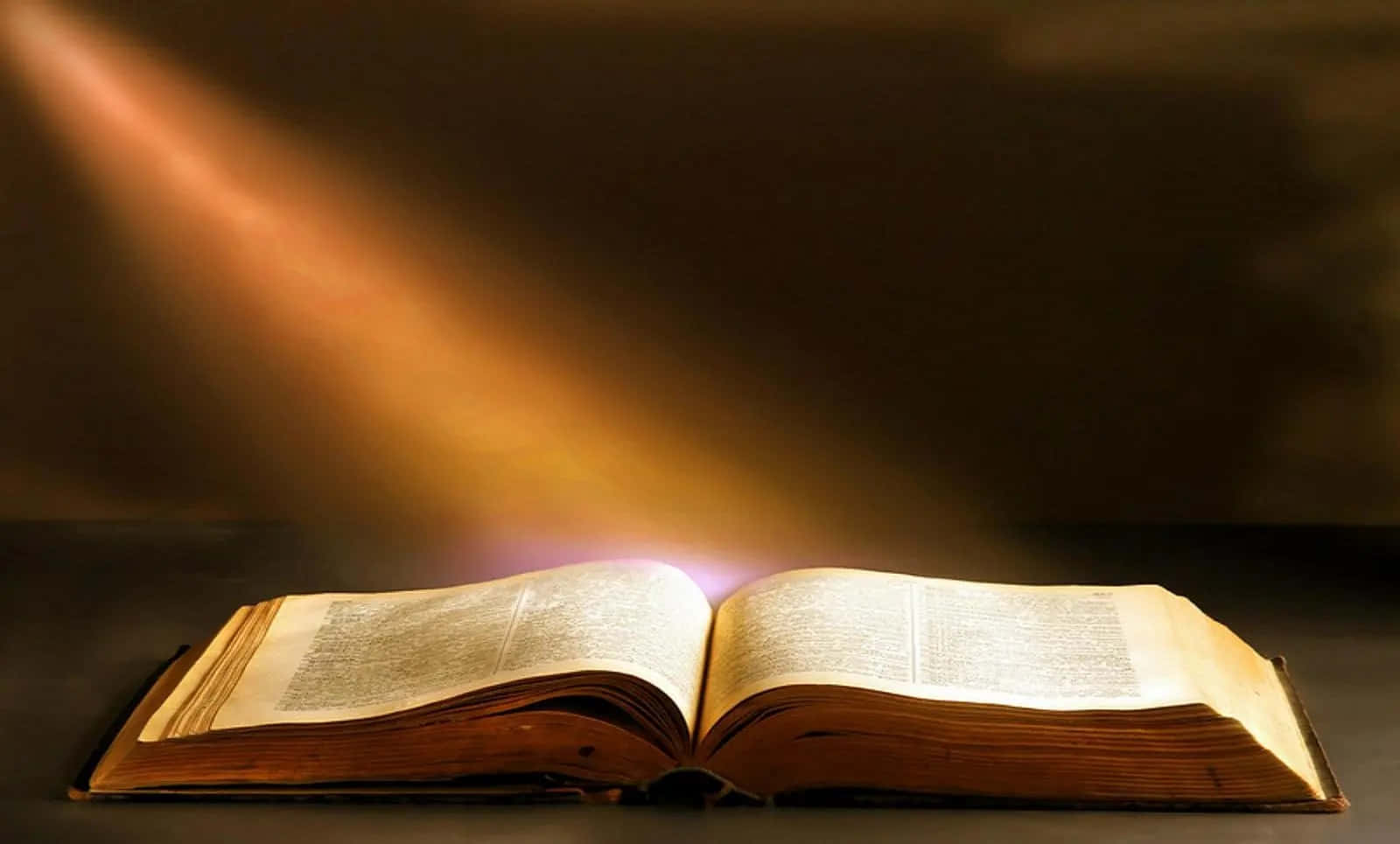 Biblical 桌布 壁紙 — 一本經風吹日曬的打開書本放在黑暗的表面上，一道溫暖的金色光束從上方戲劇性地照亮其頁面。