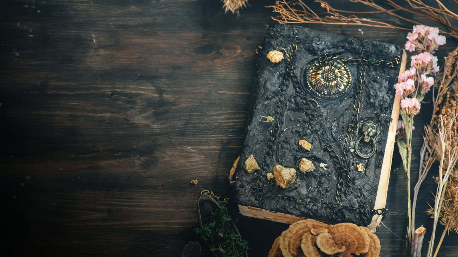 Grimoire 桌布 壁紙 — 一本華麗的黑色魔法書放在一張深色木桌上，周圍環繞著乾花和蘑菇。