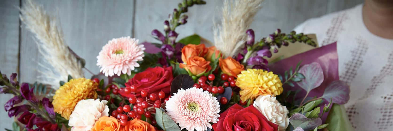 Fall Flowers 桌布 壁紙 — 一束充滿活力的秋季花束，包含紅玫瑰、橙色菊花、粉紅色非洲菊、漿果和羽毛狀的蒲葦，襯著質樸的木質背景。