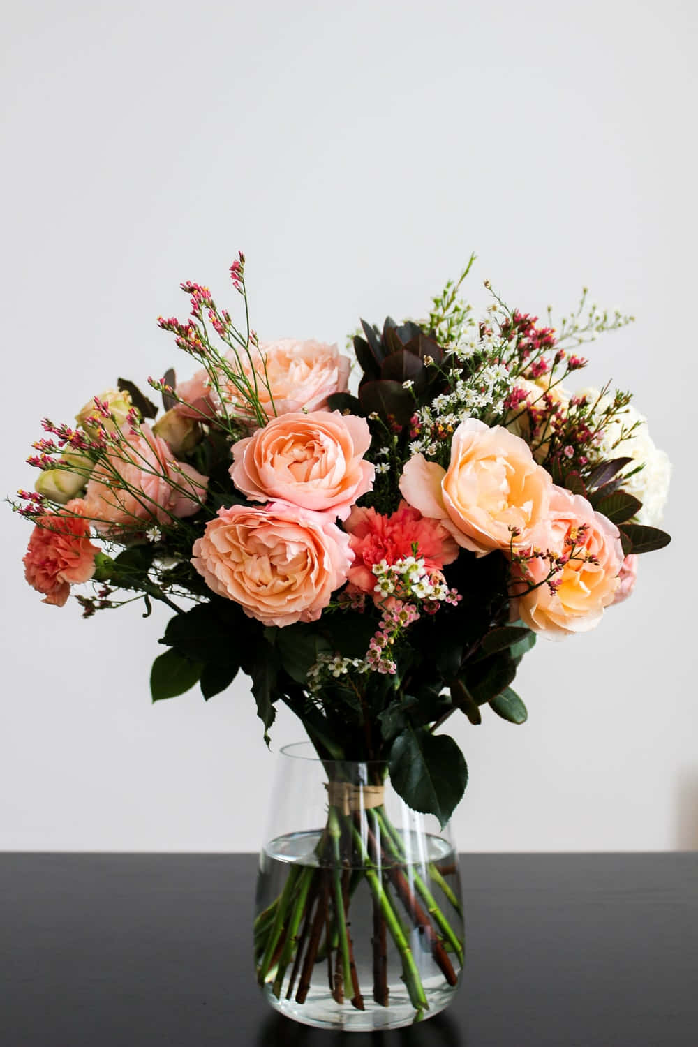 Floral Arrangement 桌布 壁紙 — 一束充滿活力的蜜桃玫瑰和精緻的填充花，放在透明玻璃花瓶裡，置於深色桌子上，背景是白色牆壁。