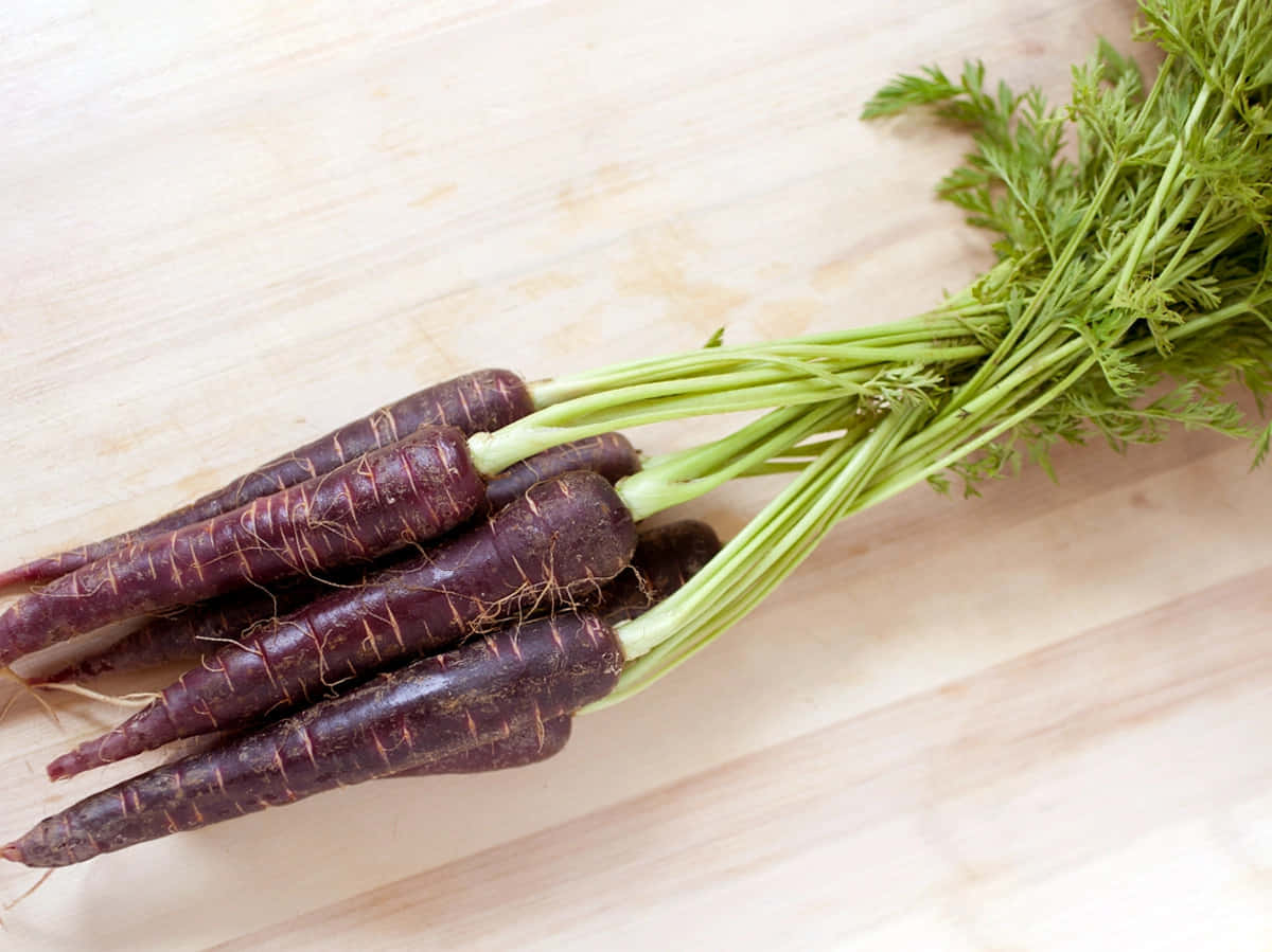 Purple Carrots 桌布 壁紙 — 一束深紫色嘅紅蘿蔔，帶有新鮮嘅綠色頂部，放喺淺色木砧板上。