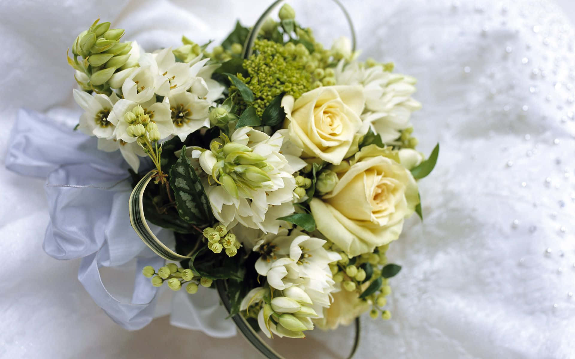 Wedding Flowers 桌布 壁紙 — 一束精緻的奶油白玫瑰和綠色植物放在透明玻璃花瓶裡，置於有紋理的白色布料上。