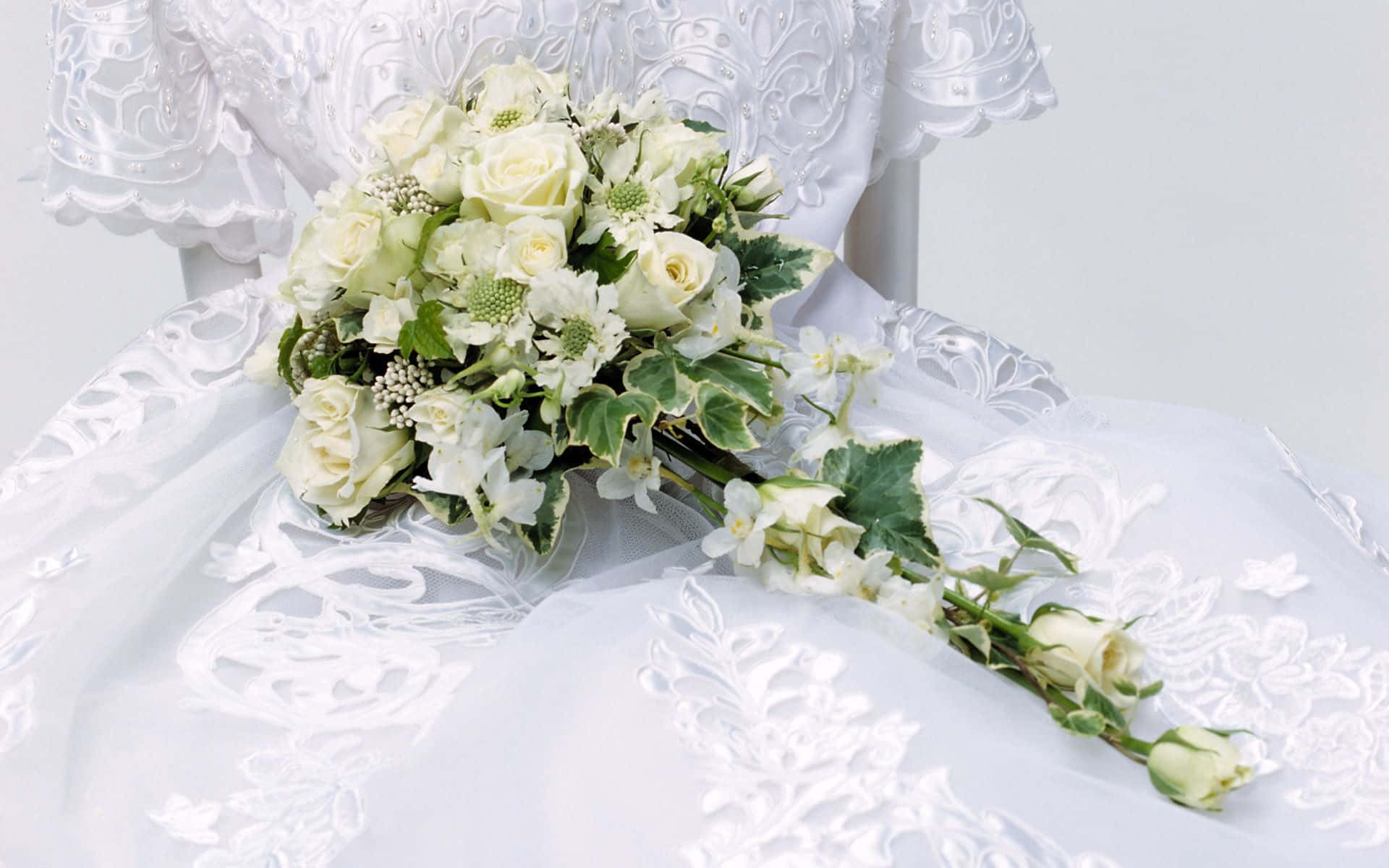 Wedding Flowers 桌布 壁紙 — 一束精緻的白色婚禮花束搭配綠色植物，放在華麗的蕾絲婚紗上，背景是柔和的白色。
