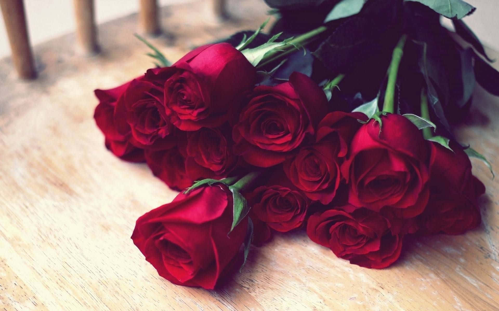 Red Rose 桌布 壁紙 — 一束茂盛的深紅玫瑰，帶有綠色莖幹，放在溫暖的木質表面上。