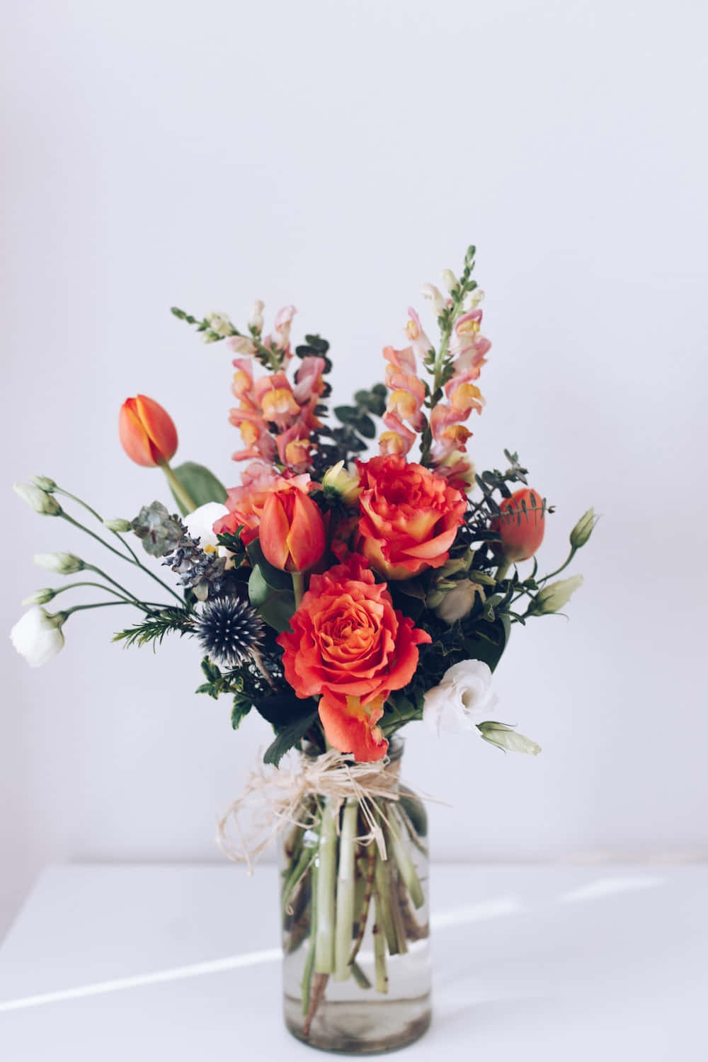 Floral Arrangement 桌布 壁紙 — 一束鮮豔的橙色玫瑰、鬱金香和金魚草，放在透明玻璃花瓶裡，用麻繩綁著，襯托簡約白色背景。