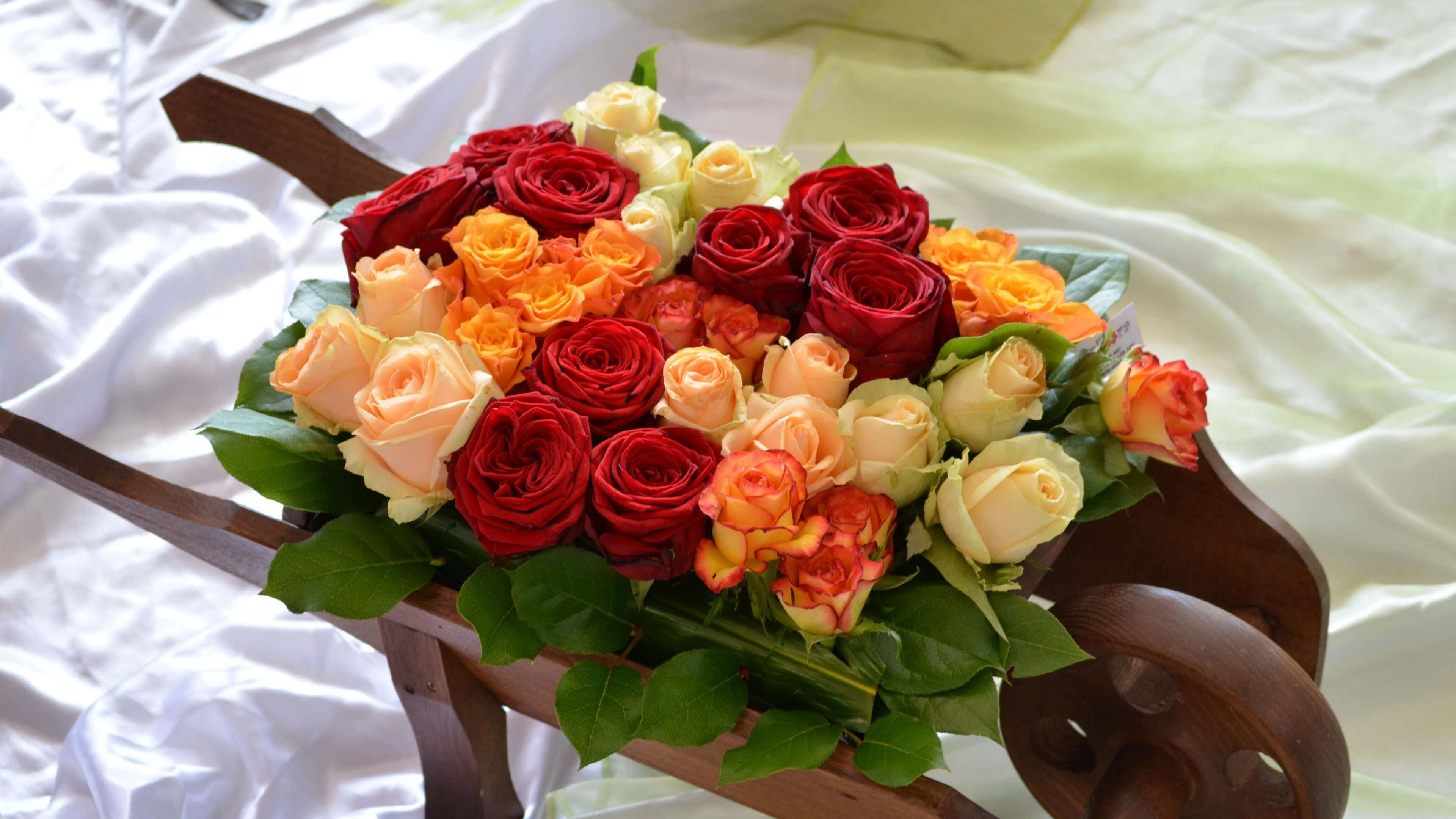 Flower Cart 桌布 壁紙 — 一束鮮豔的紅、橙和奶油色玫瑰，擺放在白色緞面床罩上的木製手推車中。