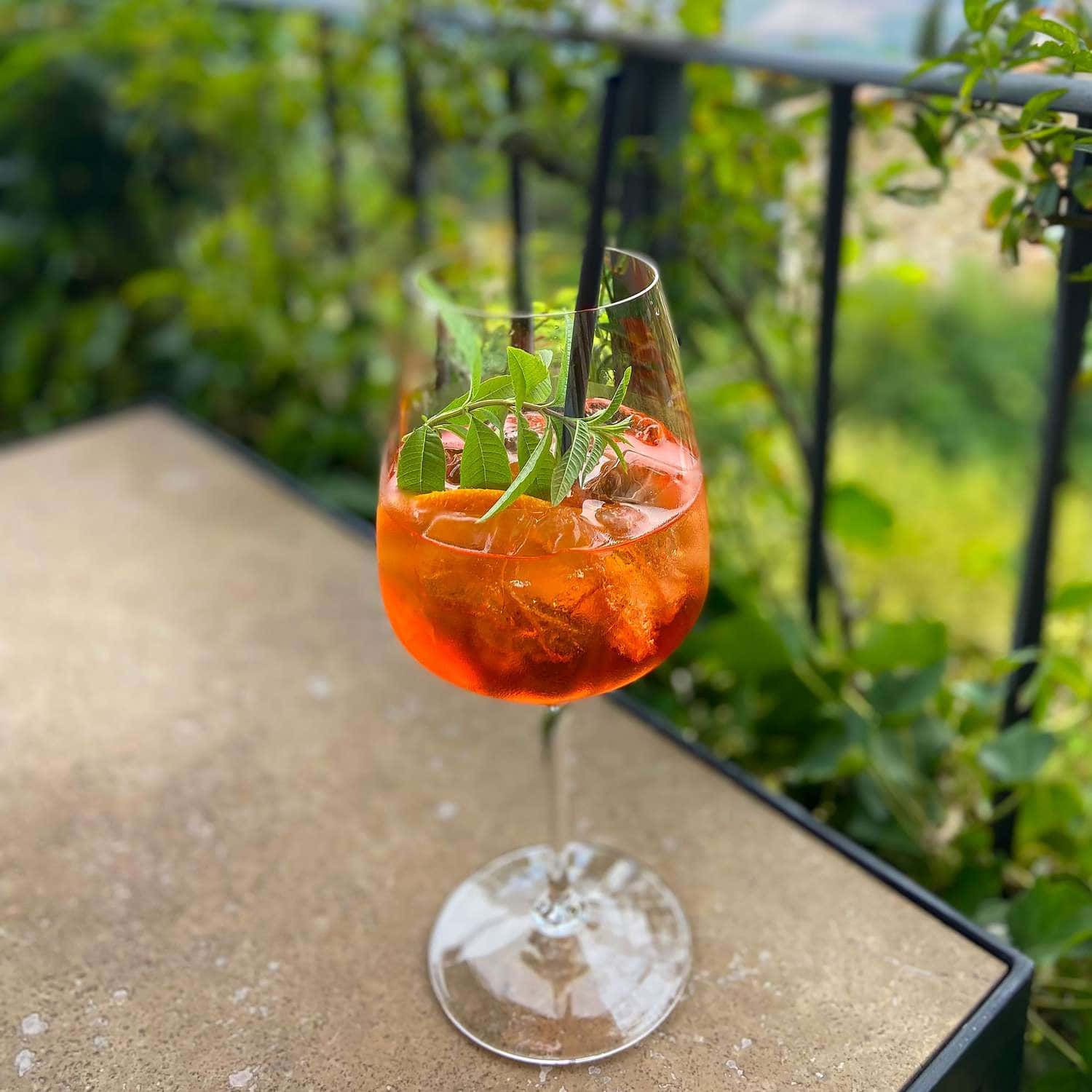 Beautiful Drink 桌布 壁紙 — 一杯充滿活力的橙色雞尾酒，加冰和新鮮香草，盛在一個高腳杯中，放在一個石頭庭院桌上，周圍環繞著茂密的綠植。