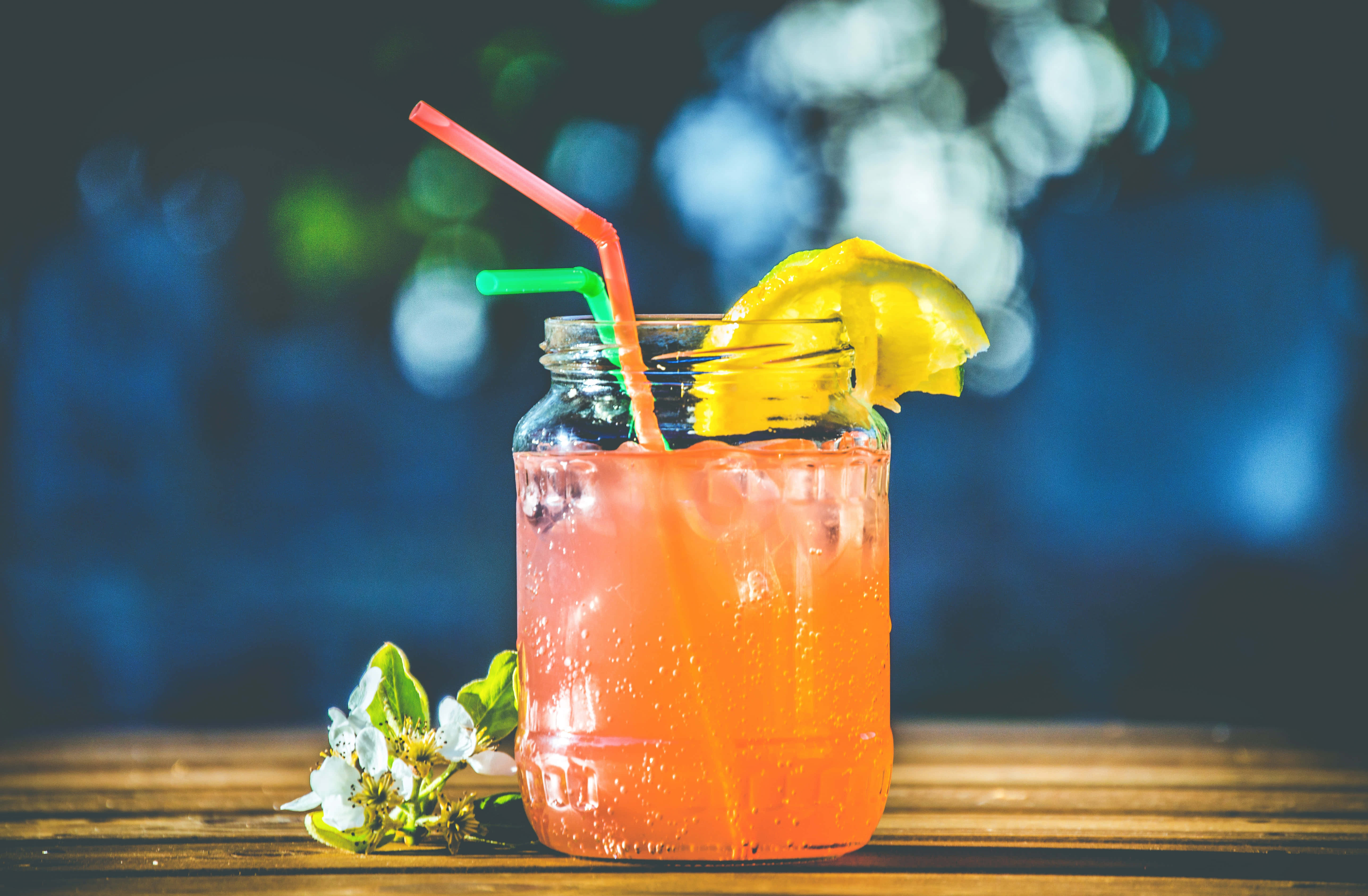 Summer Drinks 桌布 壁紙 — 一杯充滿活力的橙色飲品，配上檸檬角和兩根吸管，放在木桌上，旁邊有白色花朵，背景是模糊的藍色。