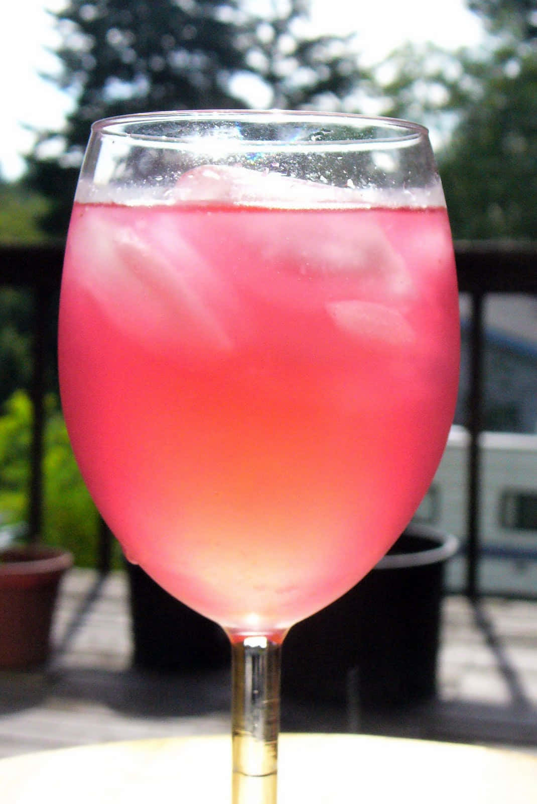 Pink Lemonade 桌布 壁紙 — 一杯充滿活力的粉紅色冰飲放在酒杯中，置於露台桌上，背景有綠植和陽光。