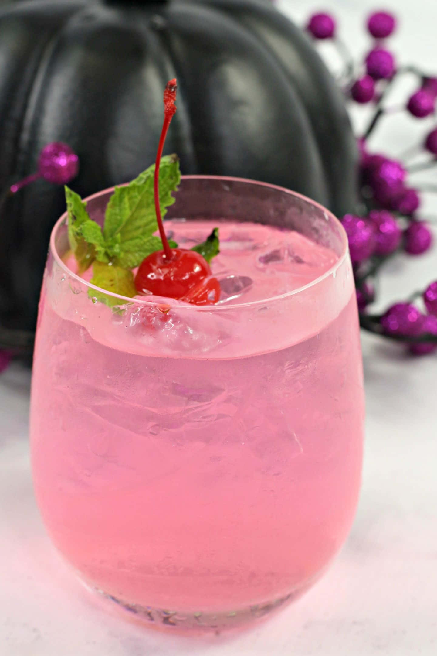Beautiful Drink 桌布 壁紙 — 一杯充滿活力的粉紅色雞尾酒，配有冰塊、櫻桃和薄荷裝飾，旁邊放著一個黑色南瓜和紫色漿果。