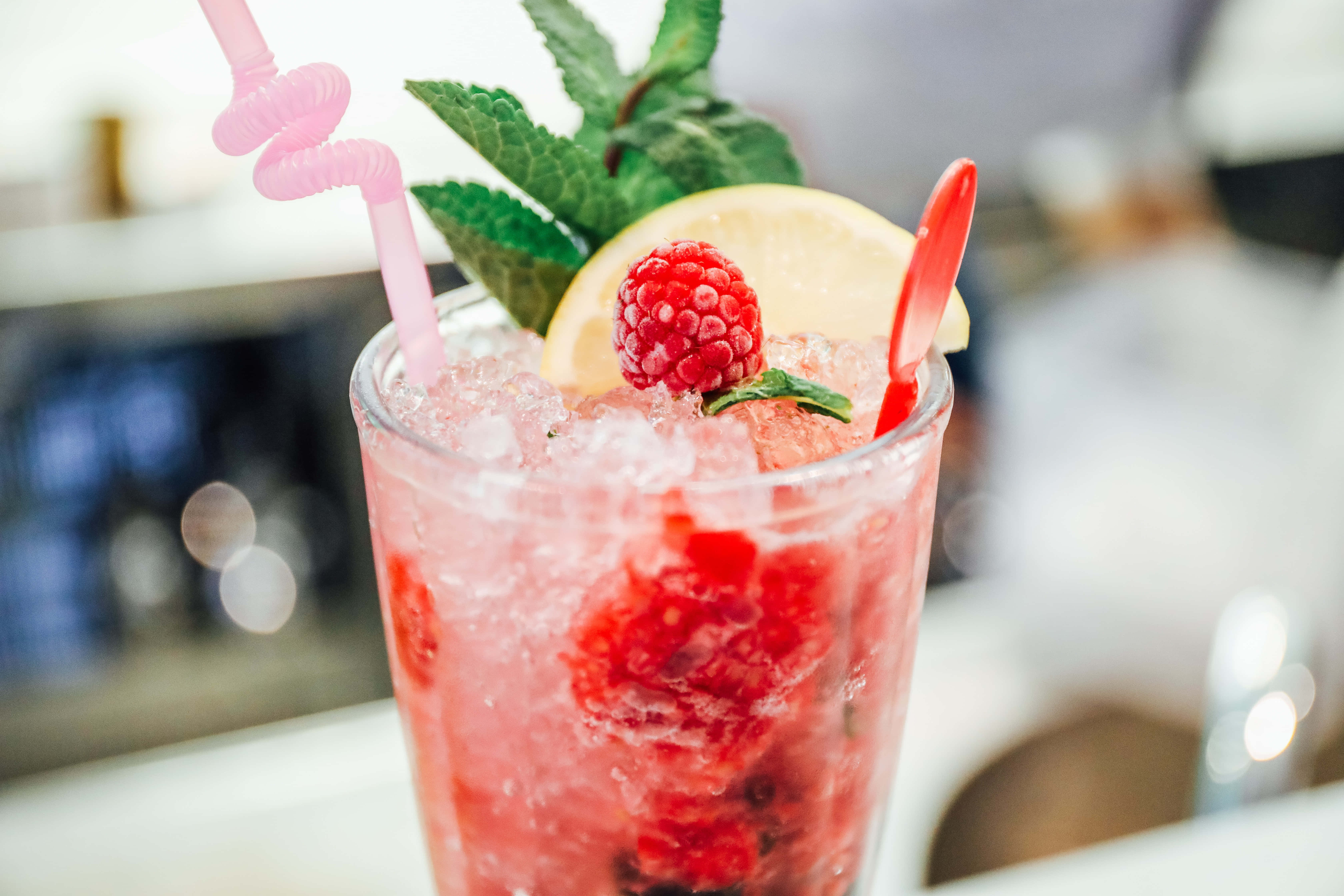 Summer Drinks 桌布 壁紙 — 一杯充滿活力的粉紅覆盆子雞尾酒，配上冰塊、薄荷、檸檬和彎曲吸管，放在酒吧枱上。