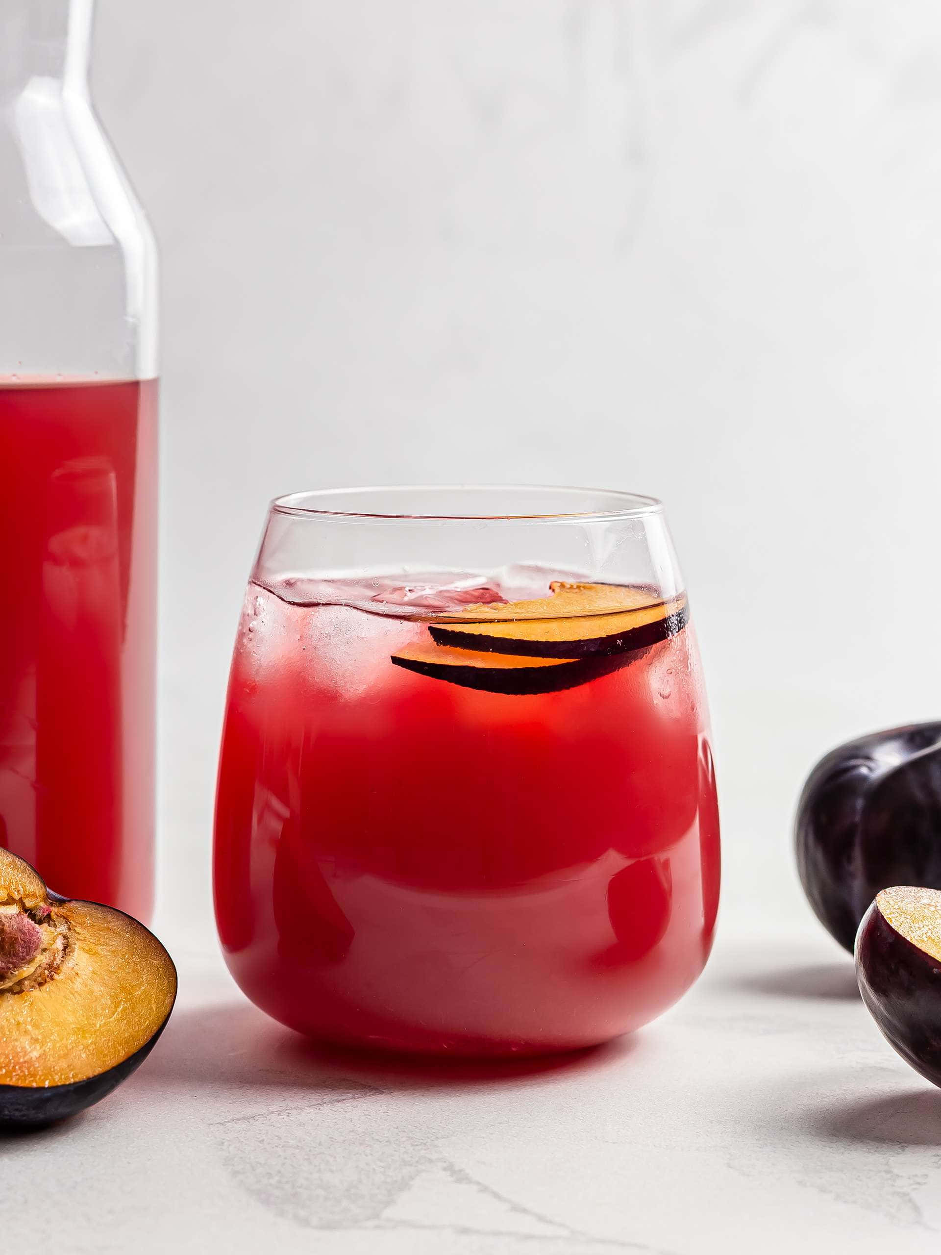Red Plum 桌布 壁紙 — 一杯充滿活力的紅梅飲品，加冰和水果片，旁邊放著一瓶酒和切半的李子，置於大理石表面上。
