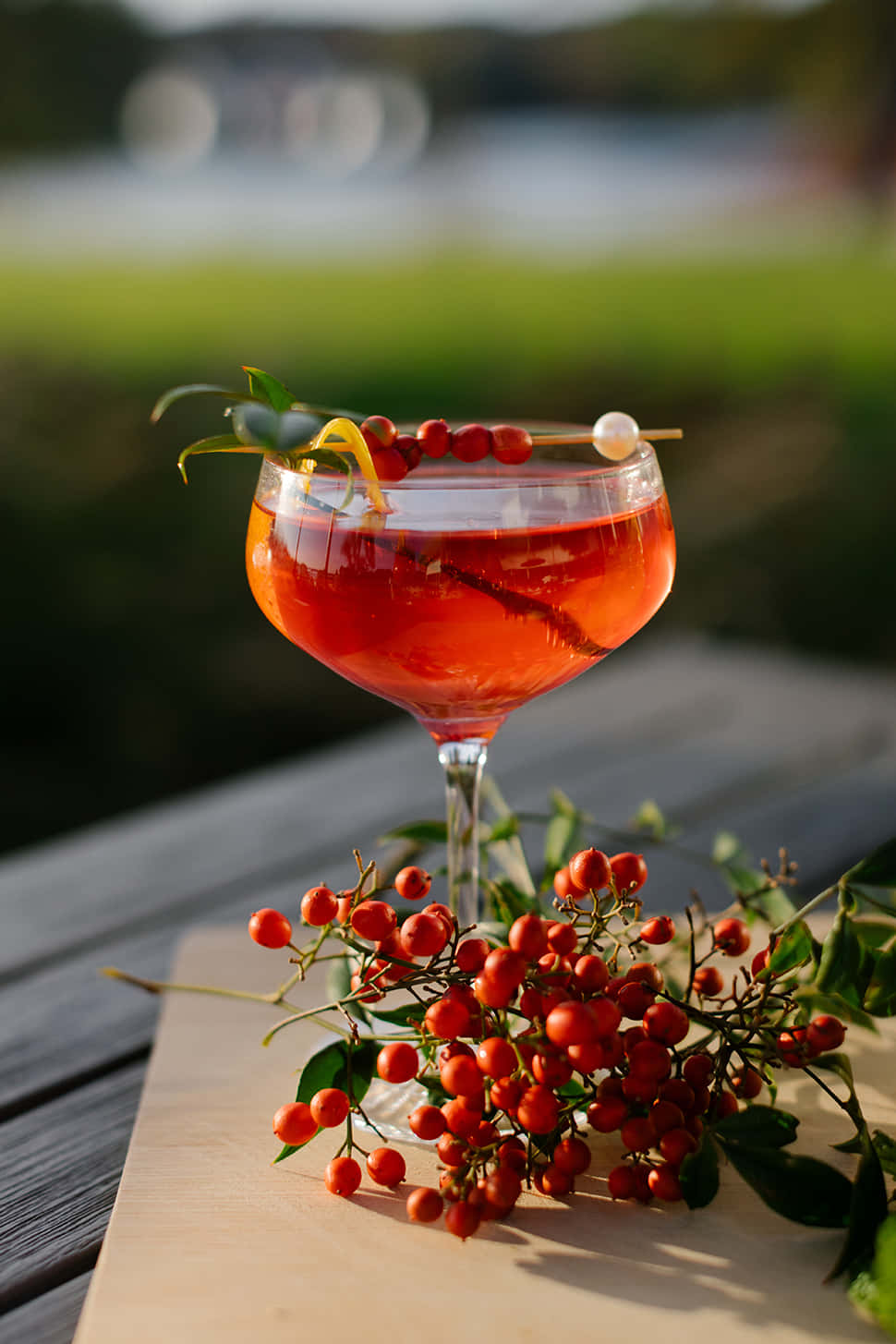 Winter Drinks 桌布 壁紙 — 一杯充滿活力的紅色雞尾酒，盛載於笛型杯中，以漿果和柑橘裝飾，旁邊放著一塊木板上盛有新鮮紅色漿果，置於陽光普照的戶外桌上。