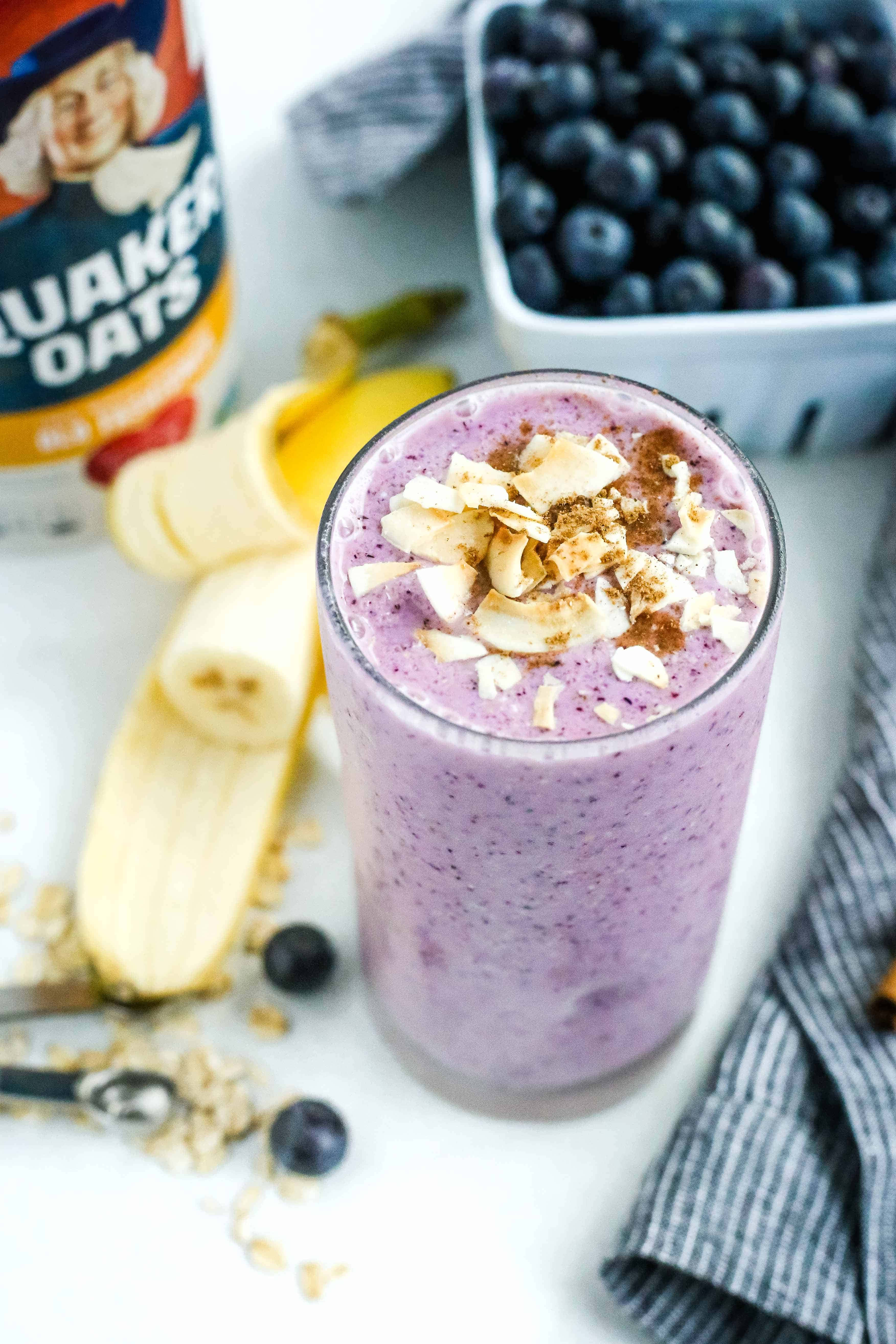 Blueberry Smoothie 桌布 壁紙 — 一杯充滿活力的紫色冰沙，上面撒上椰子片和肉桂，旁邊是新鮮藍莓、香蕉和Quaker燕麥片，放在白色表面上。